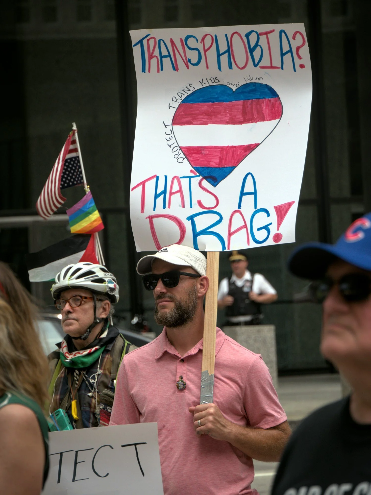   A man in a pink polo, white baseball cap, and sunglasses holds a protest sign that reads 