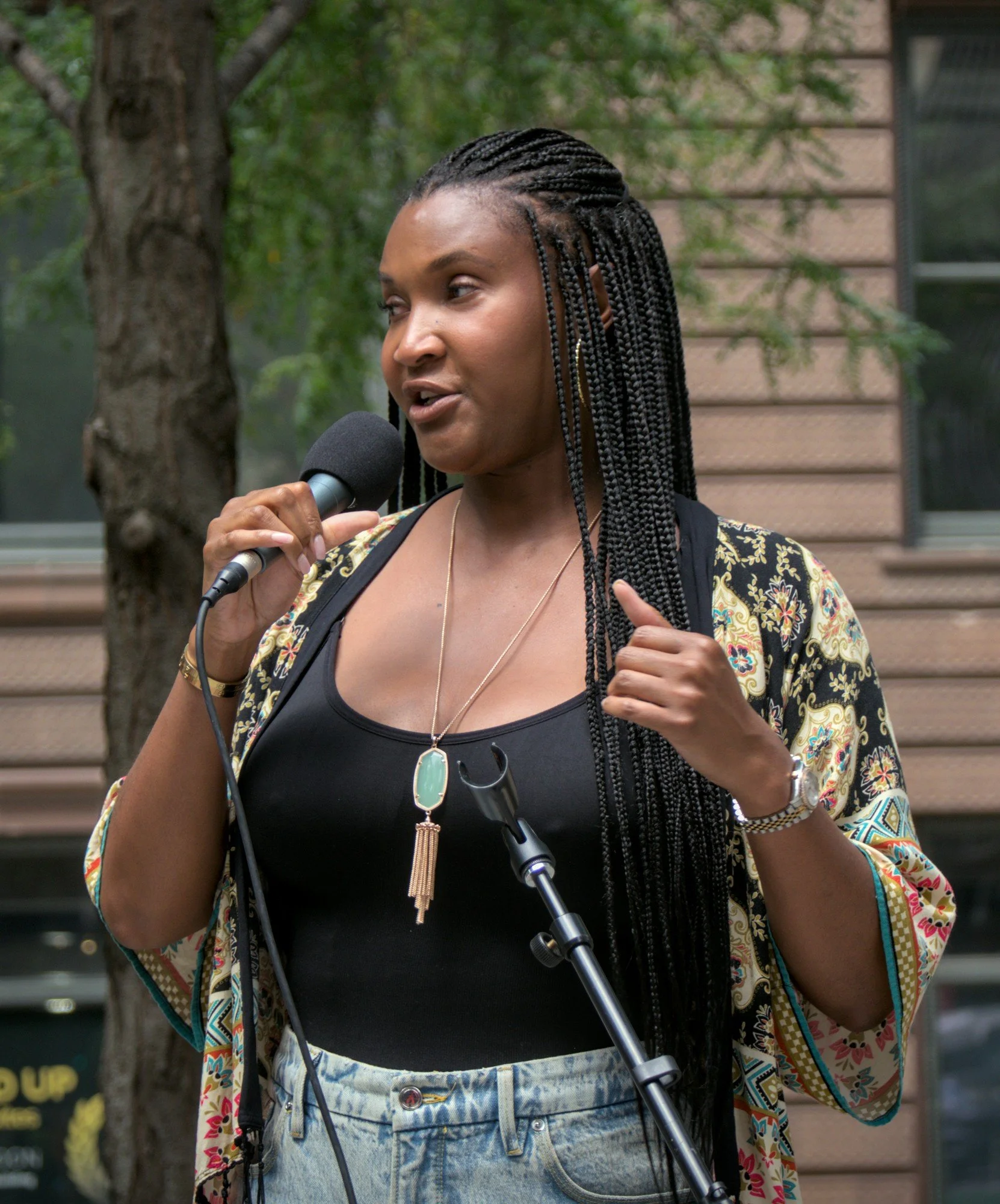   Channyn Lynne Parker, sporting long black braids and a long necklace and wearing a silk topper, a black camisole, and blue jean shorts, speaks to the crowd.  