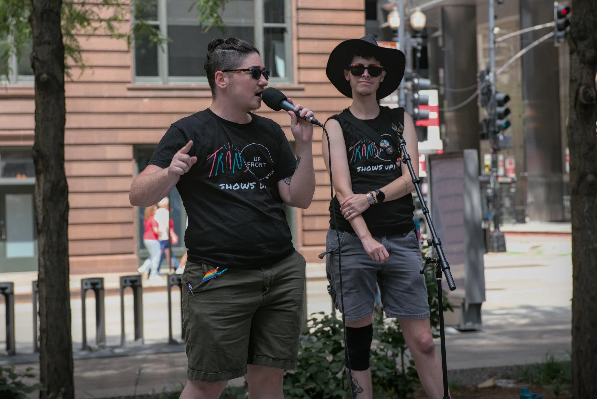   A portrait of Asher Mcmaher--on the left, with short cropped hair tied back in a bun and wearing sunglasses and green cargo shorts-- and co-founder Charlee Friedman -wearing grey cargo shorts and a sun hat--at the microphone.  