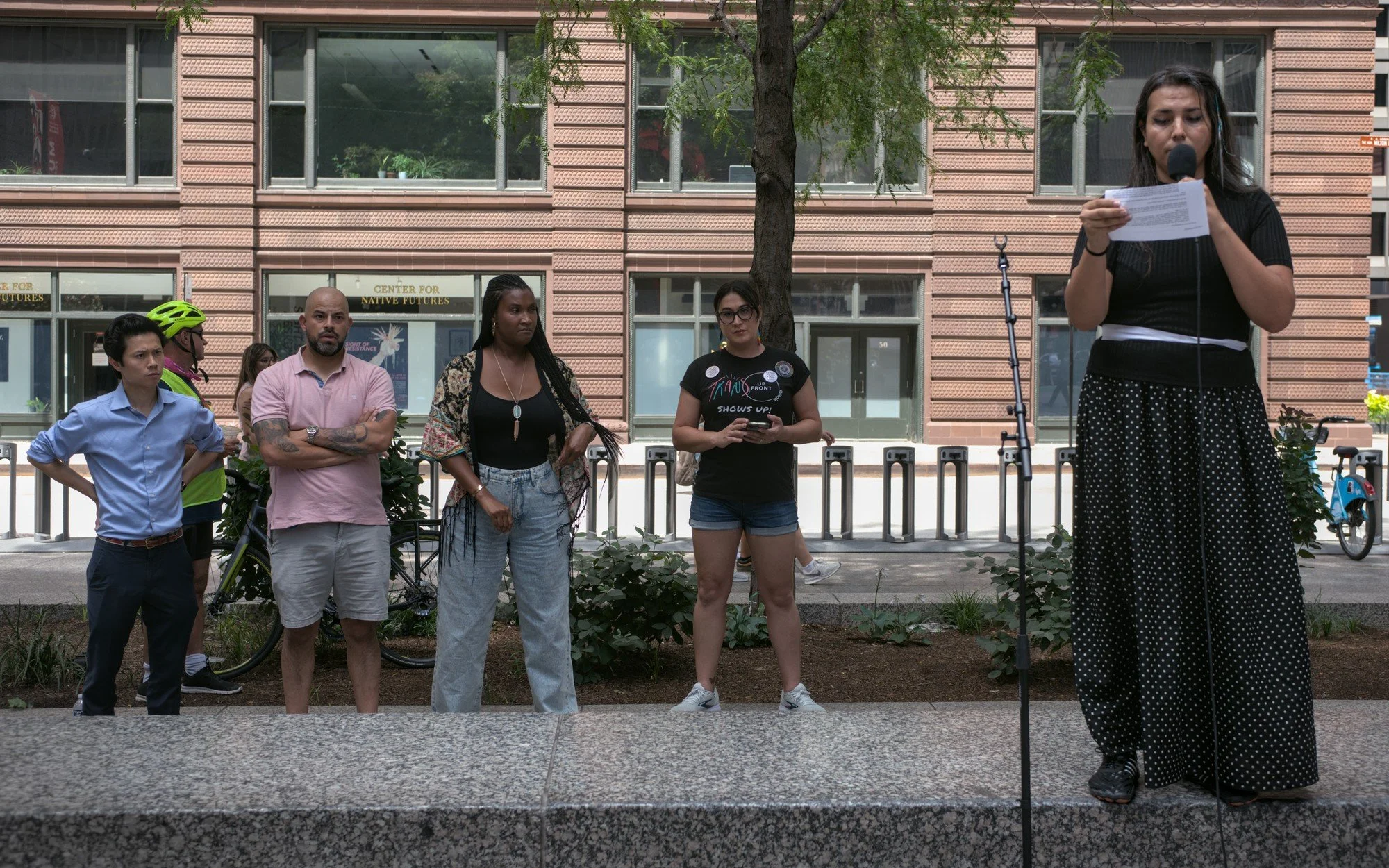   Albie Gutierrez, a brown-haired femme with a blue beaded streak tied into her hair wearing a skirt and black shirt, stands at the far right of the image. Four of the other speakers--Hoan Huynh, Andre Vasquez, Channyn Lynne Parker and Corey Lascano--stand in a line behind the marble bench the protesters are using as a makeshift stage.  