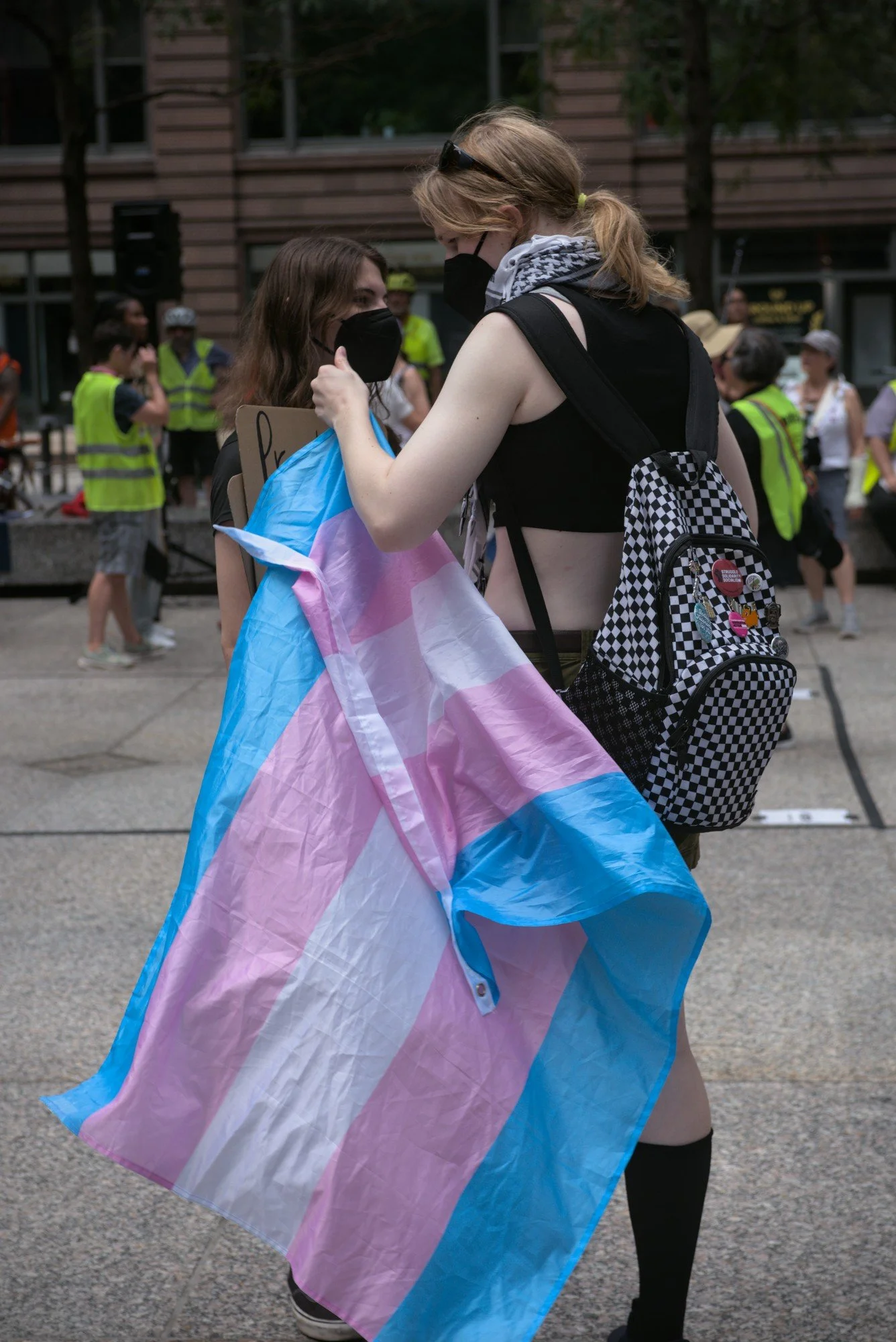   Two trans femmes stand in the plaza, one of which unravels a trans pride flag she was wearing as a cape. A group of yellow-vested folk, most likely volunteers for Indivisible, pepper the background.  