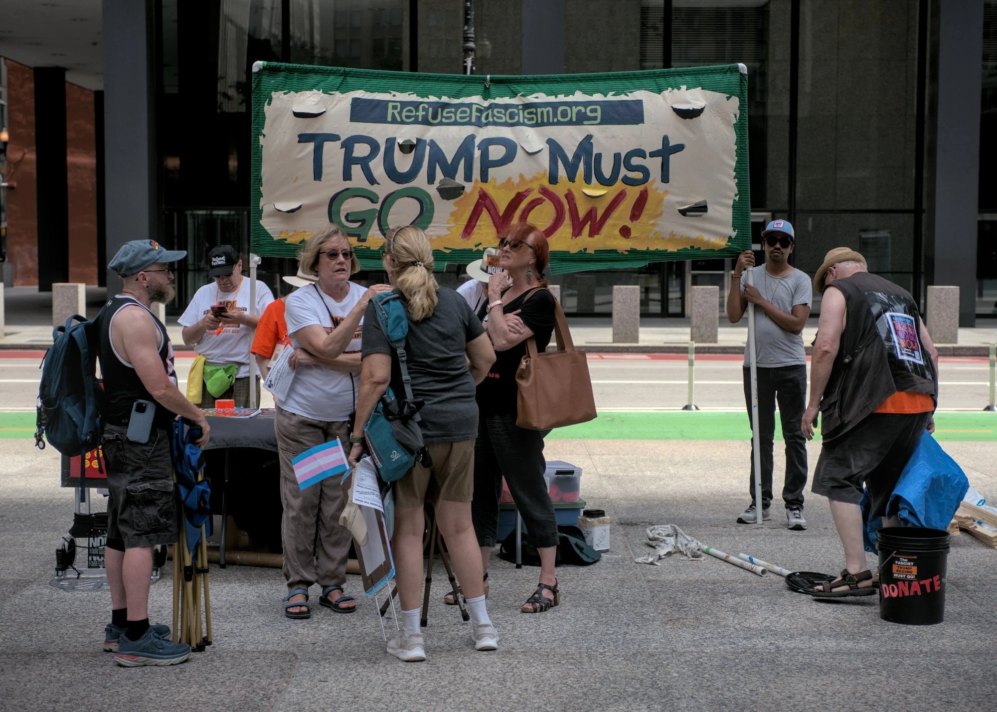   Another group sets up near the protest, this one for    Refusefascism.org   . A small gathering of older white femmes talk and commiserate about the current state of affairs in the foreground, while a masc in a tank and shorts patiently waits to talk to one of them and a man in a blue hat holds the banner awkwardly in the background while staring at the viewer.  