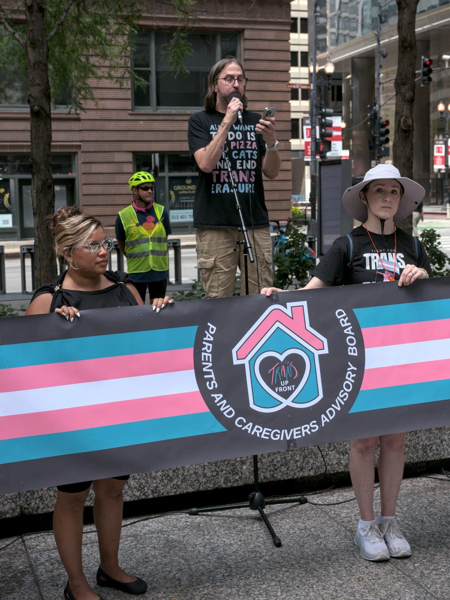   The Parents And Caregivers Advisory Board for Trans Up Front holds a long trans pride flag banner, with speaker Bob—a long-haired masc figure wearing glasses, sporting long sandy blonde hair, and with a shirt that says ALL I WANT TO DO IS EAT PIZZA PET CATS AND END TRANS ERASURE—behind the banner, reading a prepared speech to the crowd.  