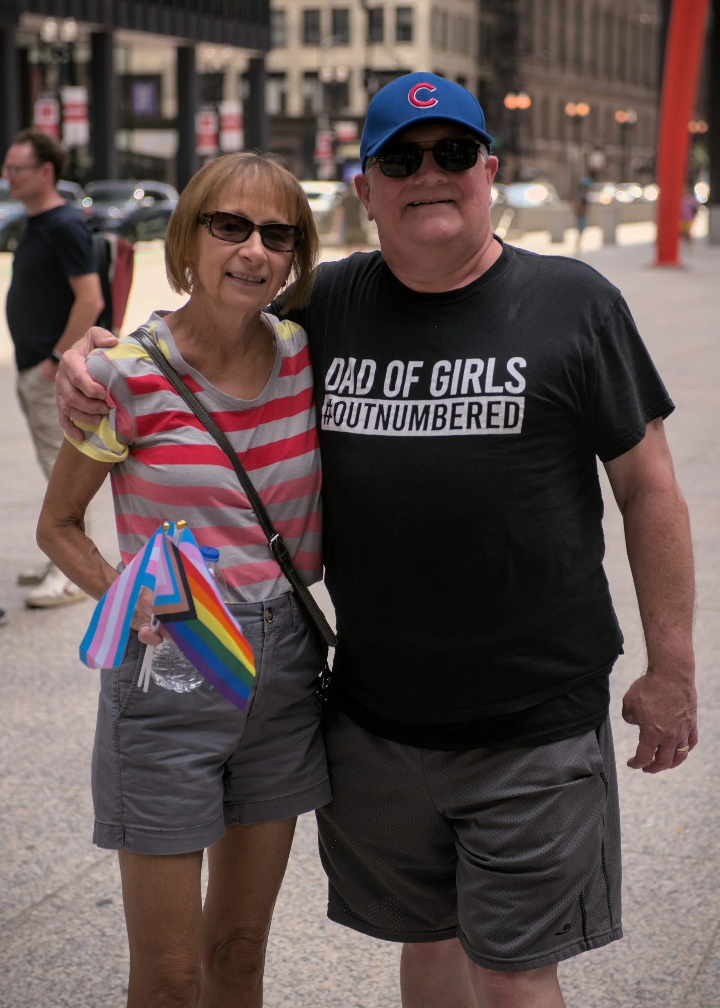   A portrait of Ed and Tracy Dembek. Tracy is on the left, wearing sunglasses and a striped shirt and carrying a Progress pride flag and a trans flag. Ed is on the right, wearing a shirt which says 