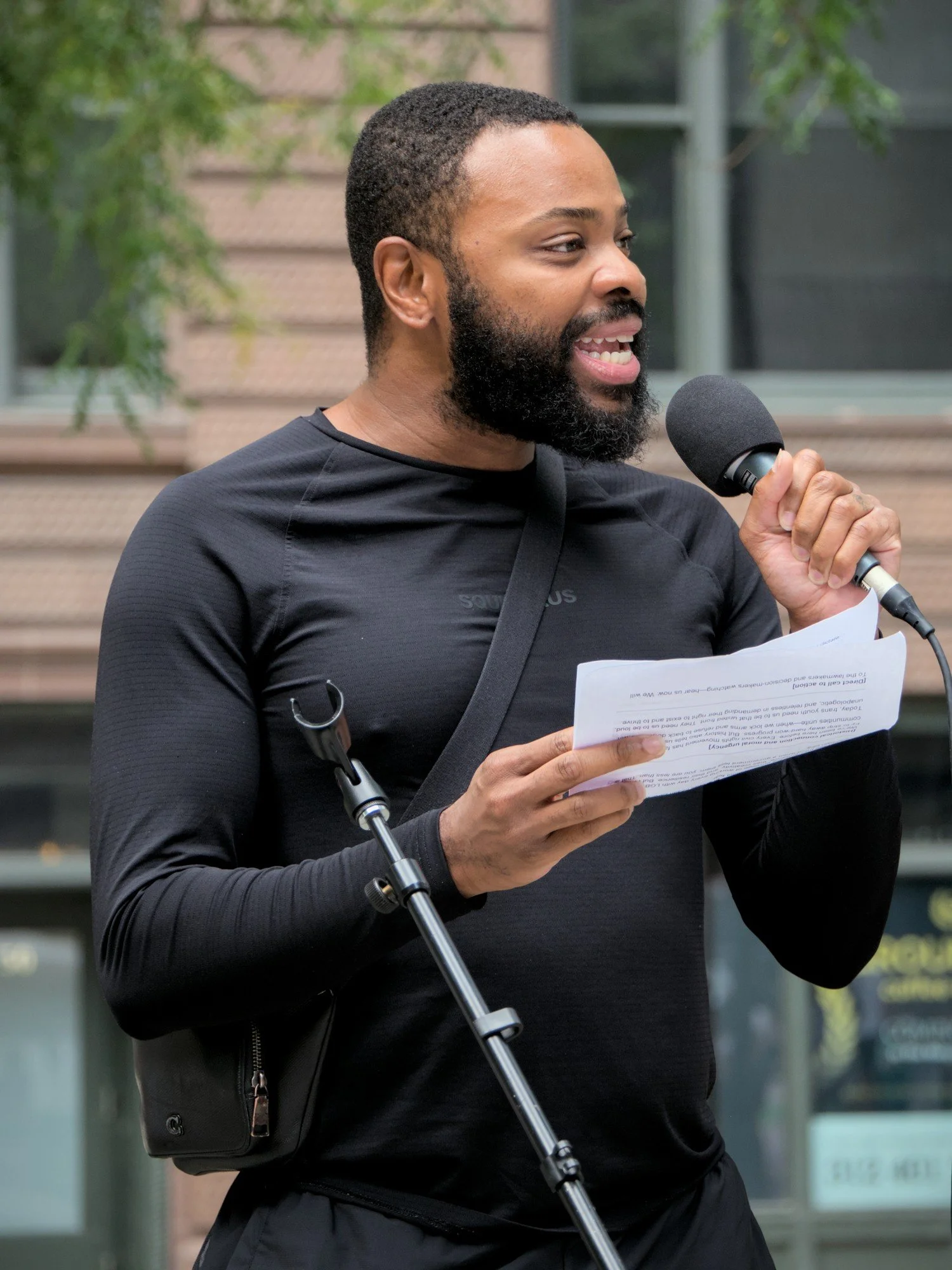   Dr. Chris Balthazar, executive director of Task Force Chicago  and sporting a beard and short cropped hair and wearing a black workout shirt and shorts, speaks to a crowd into a microphone, reading off of a note-sheet.  