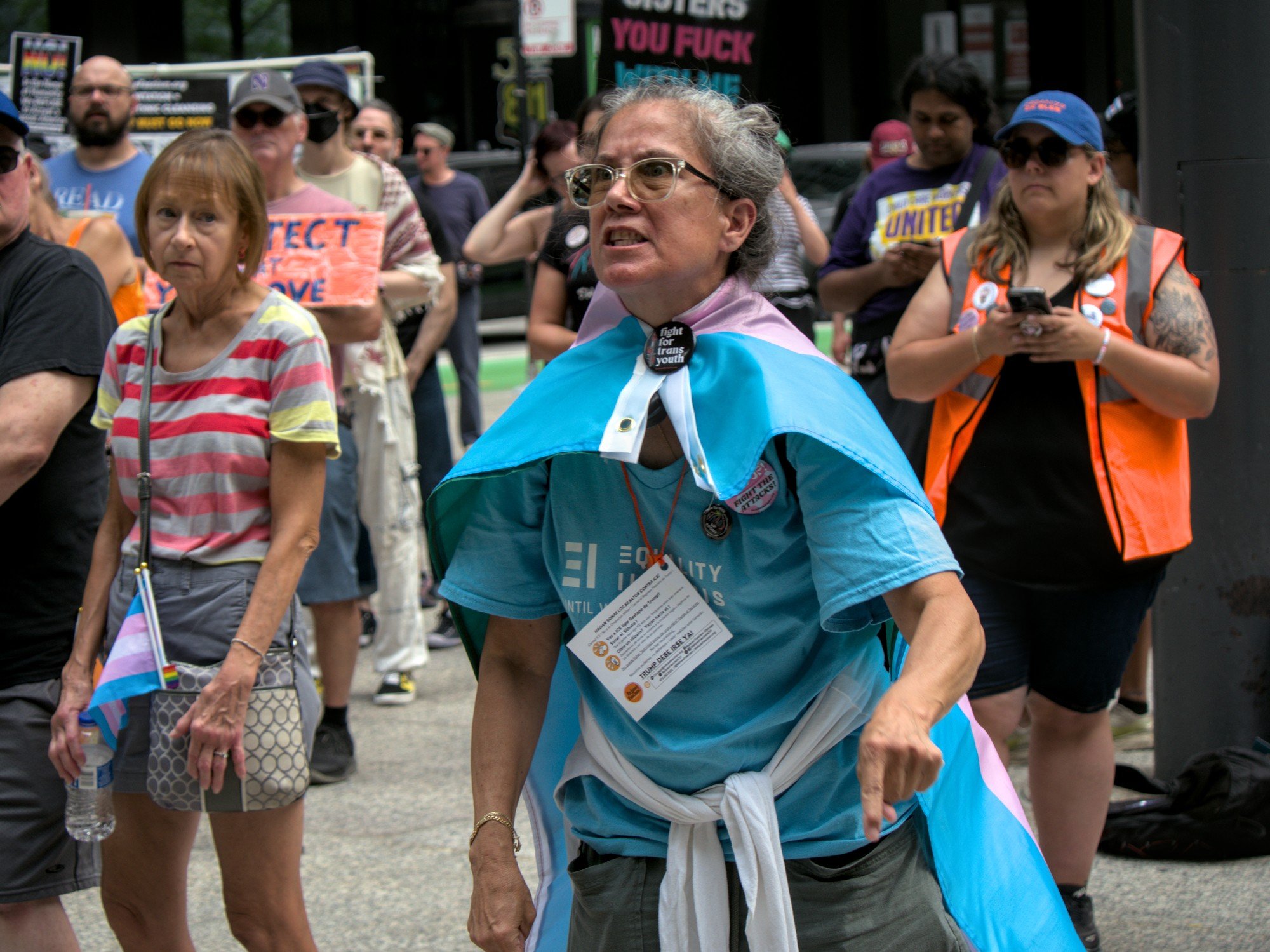   A woman in a blue shirt and glasses with a trans flag wrapped around her as a cape gestures angrily towards someone offscreen, while members of the crowd look on.  