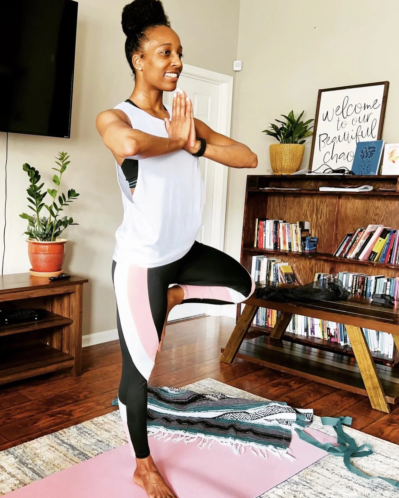 Woman practicing yoga indoors in a tree pose, standing on a pink yoga mat, with a bookshelf, potted plants, and a framed sign that reads 'welcome to our beautiful chaos' in the background.