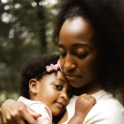 A woman hugging a young girl outdoors in a forest setting.