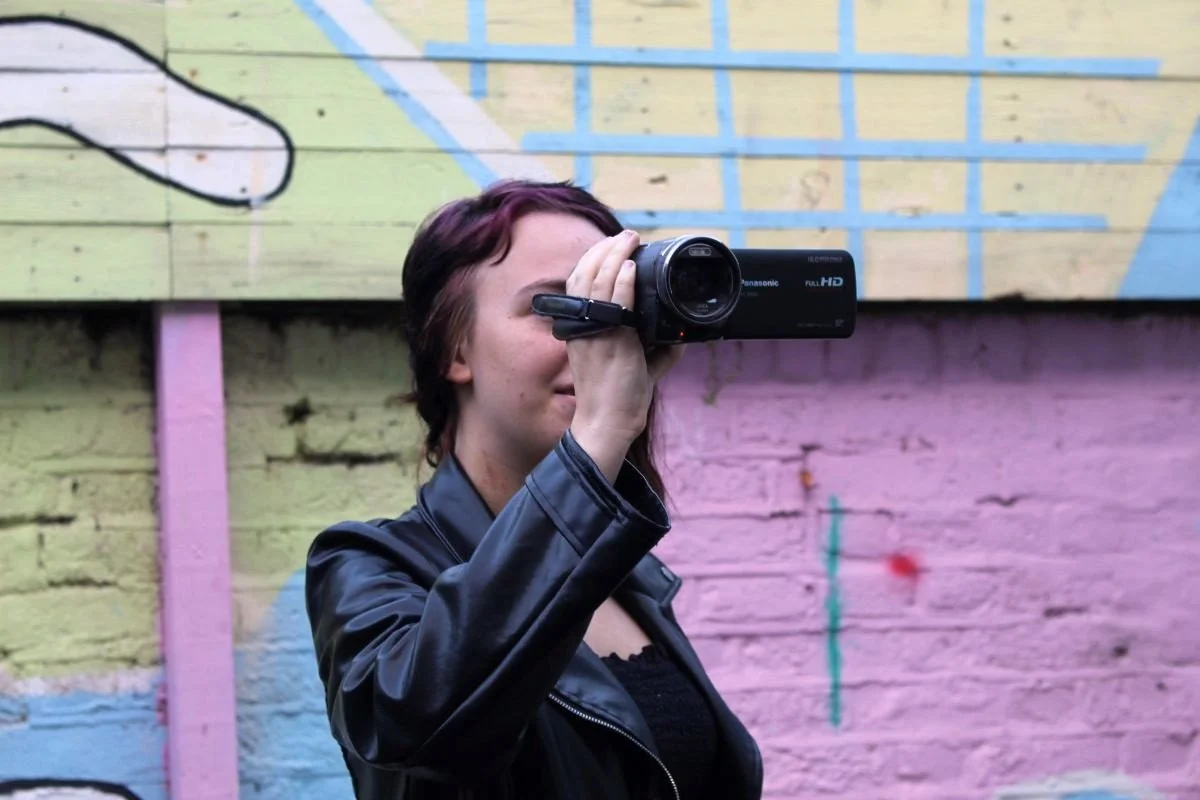 A young person recording with a video camera in our courtyard. Thye are stood in front of our colourful pastel mural.