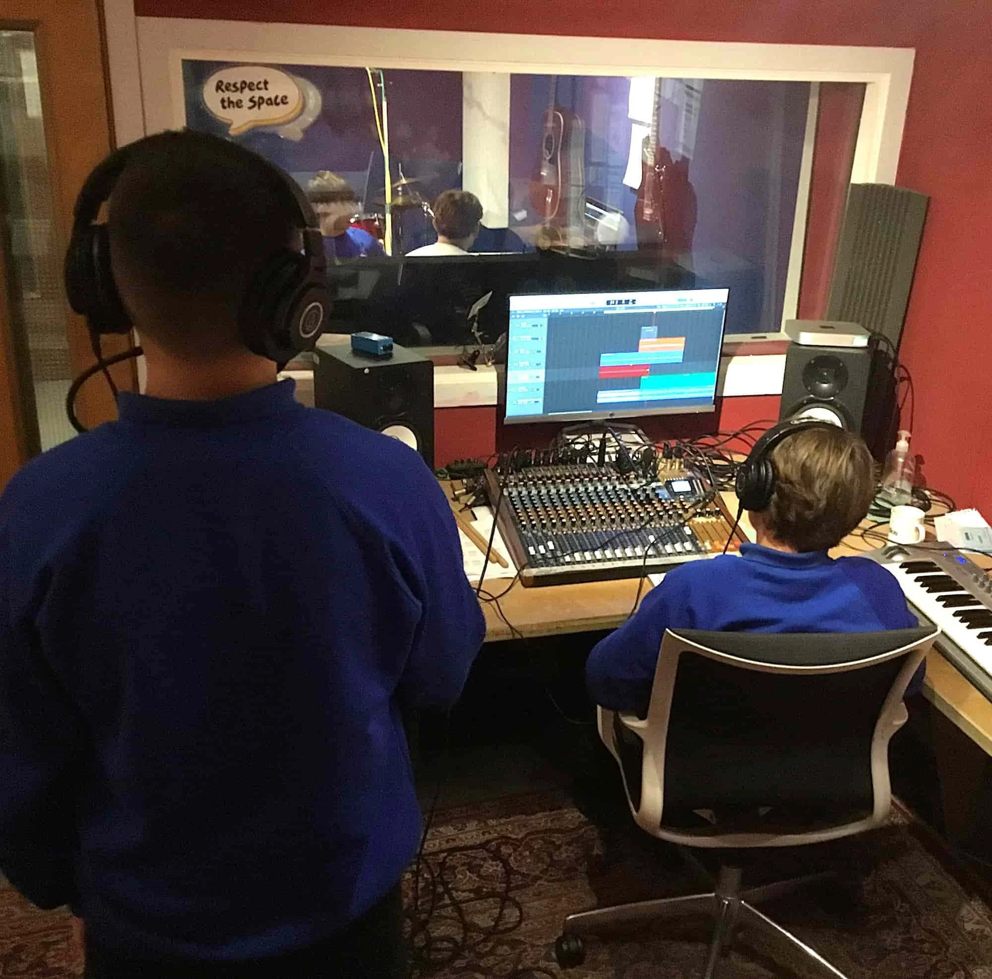 Two young people with headphones on making music, one is sat at the mixing desk and the other is stood in front of the microphone..