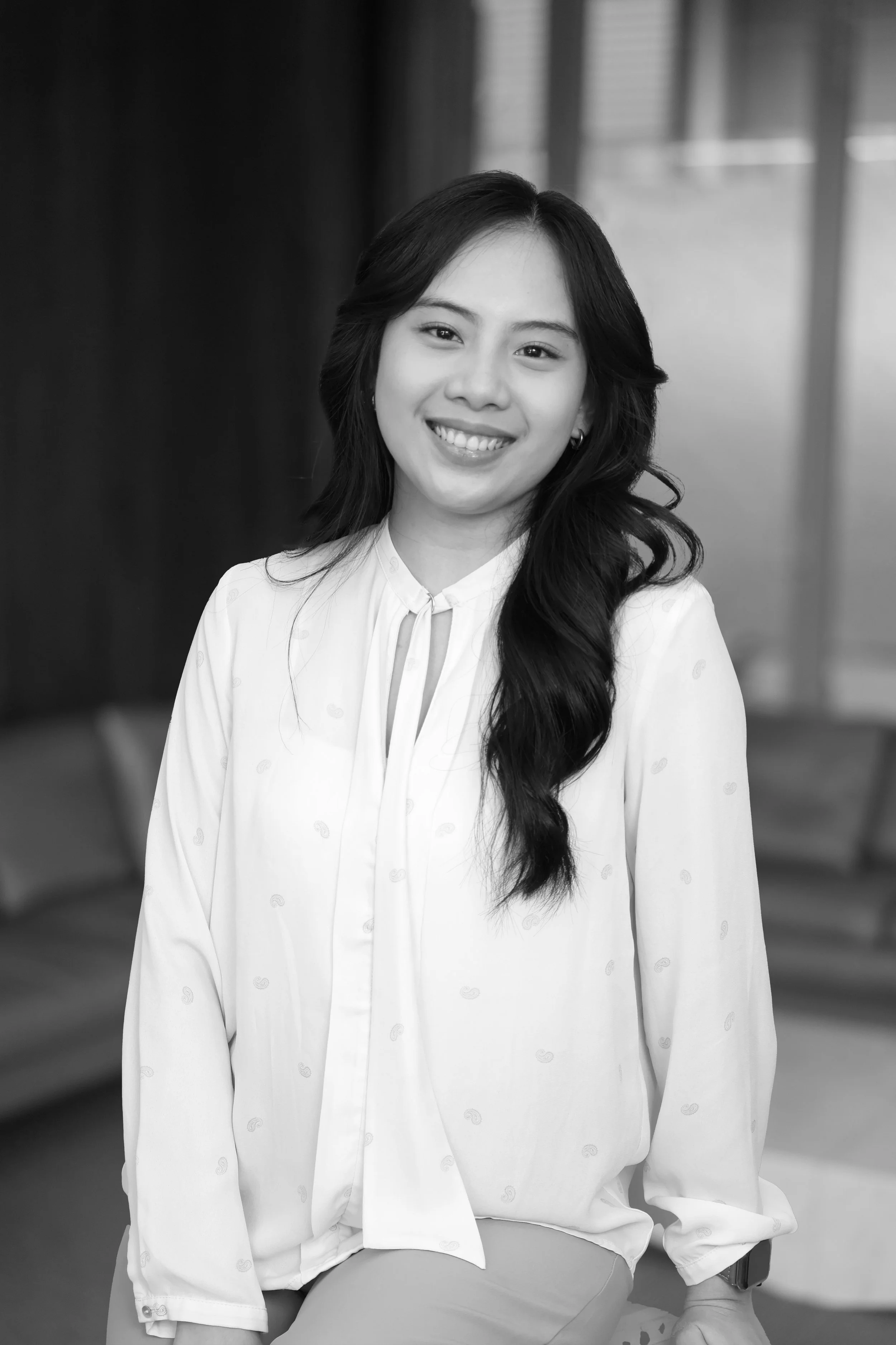 A smiling young woman with long, wavy hair in a professional or casual setting, wearing a light-colored blouse. The background features a room with seating and large windows.