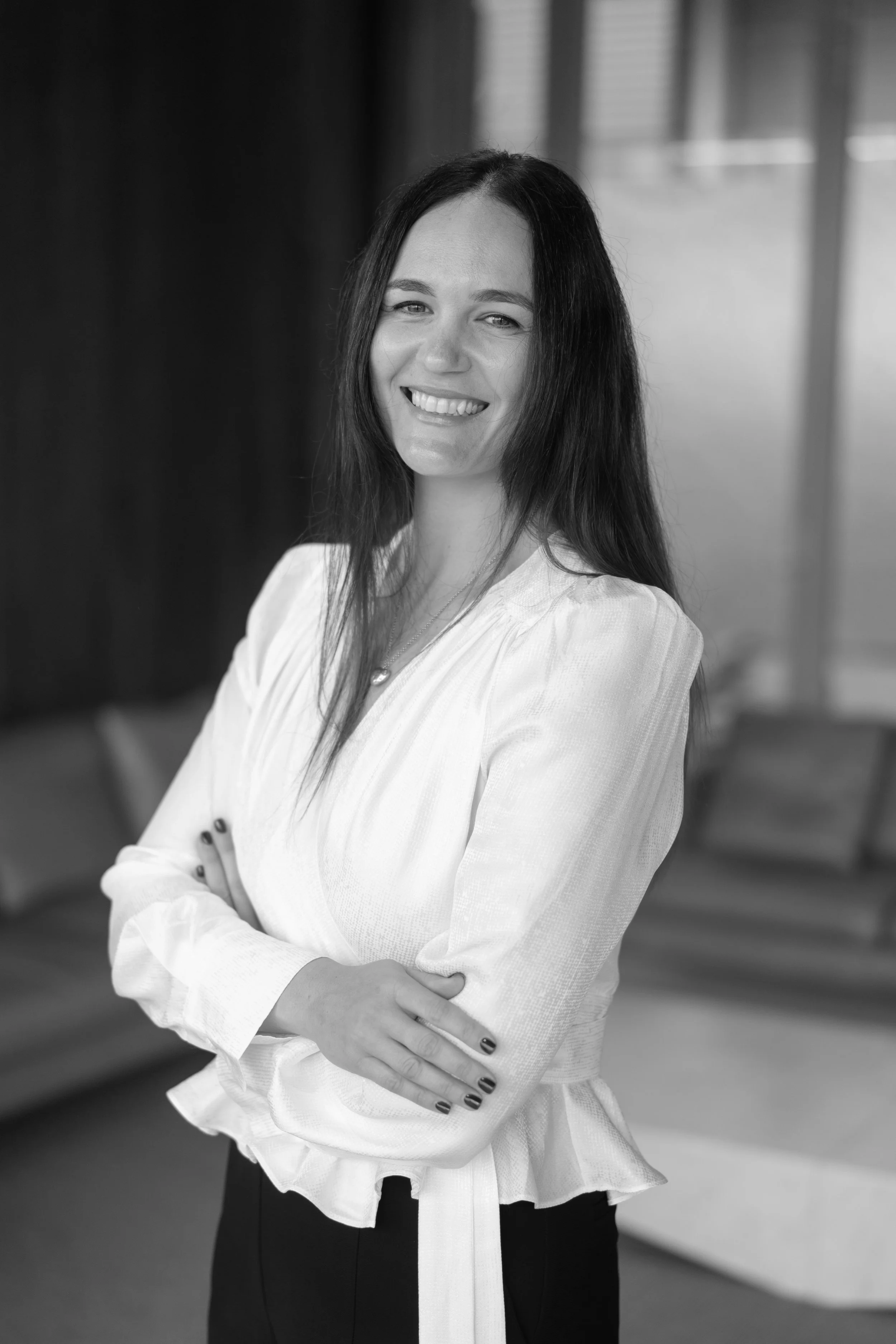 A smiling woman with long dark hair wearing a light-colored blouse and dark pants, standing with her arms crossed in an indoor setting.