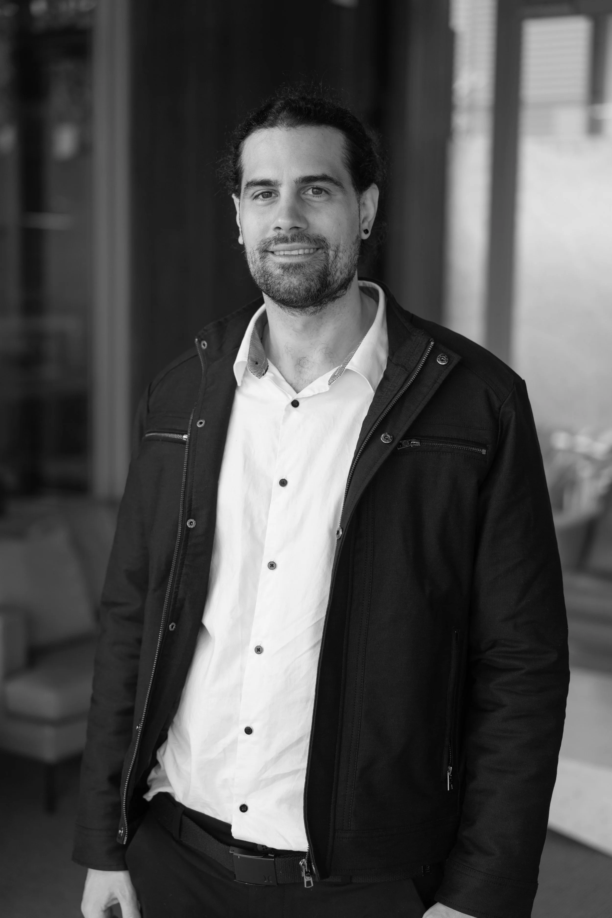 Black and white portrait of a man with dark curly hair, beard, and earrings, wearing a white dress shirt and a dark jacket, standing indoors with a blurred background.