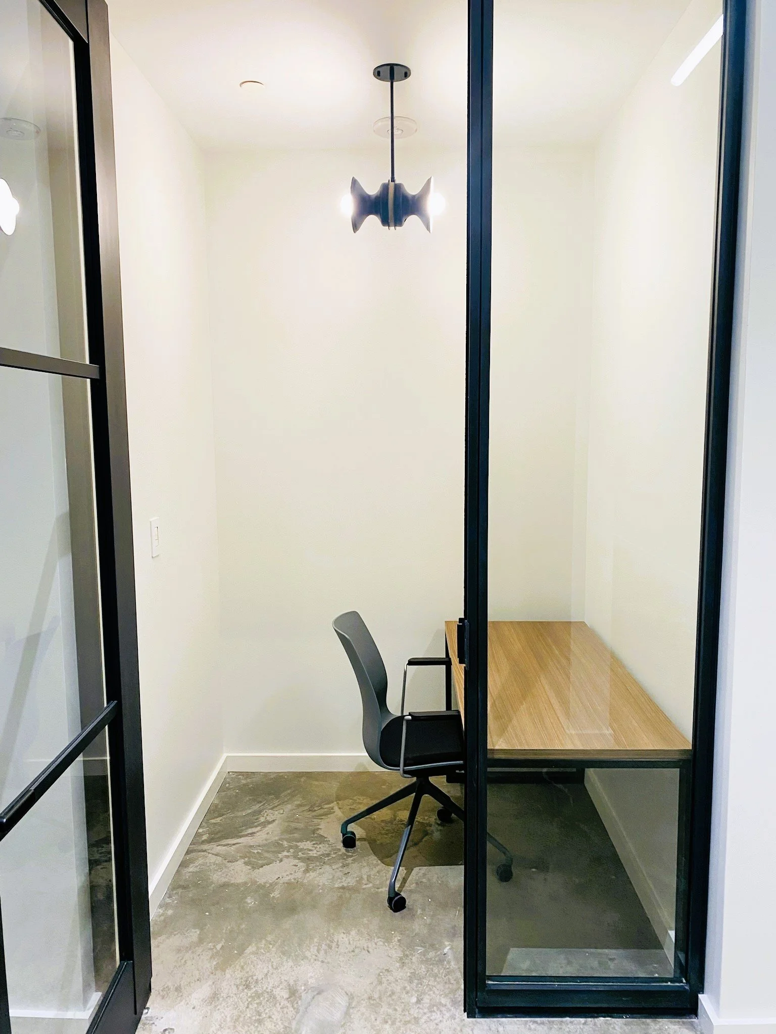 Empty modern office cubicle with black-framed glass walls, a black office chair, a small wooden desk, and a black hanging light fixture, with concrete floor and white walls.