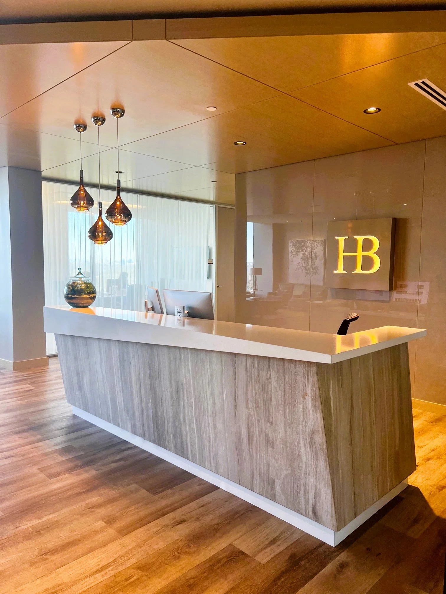 Modern hotel reception desk with a white marble countertop, wooden paneling, decorative lighting, and a backlit 'HB' sign on the wall.