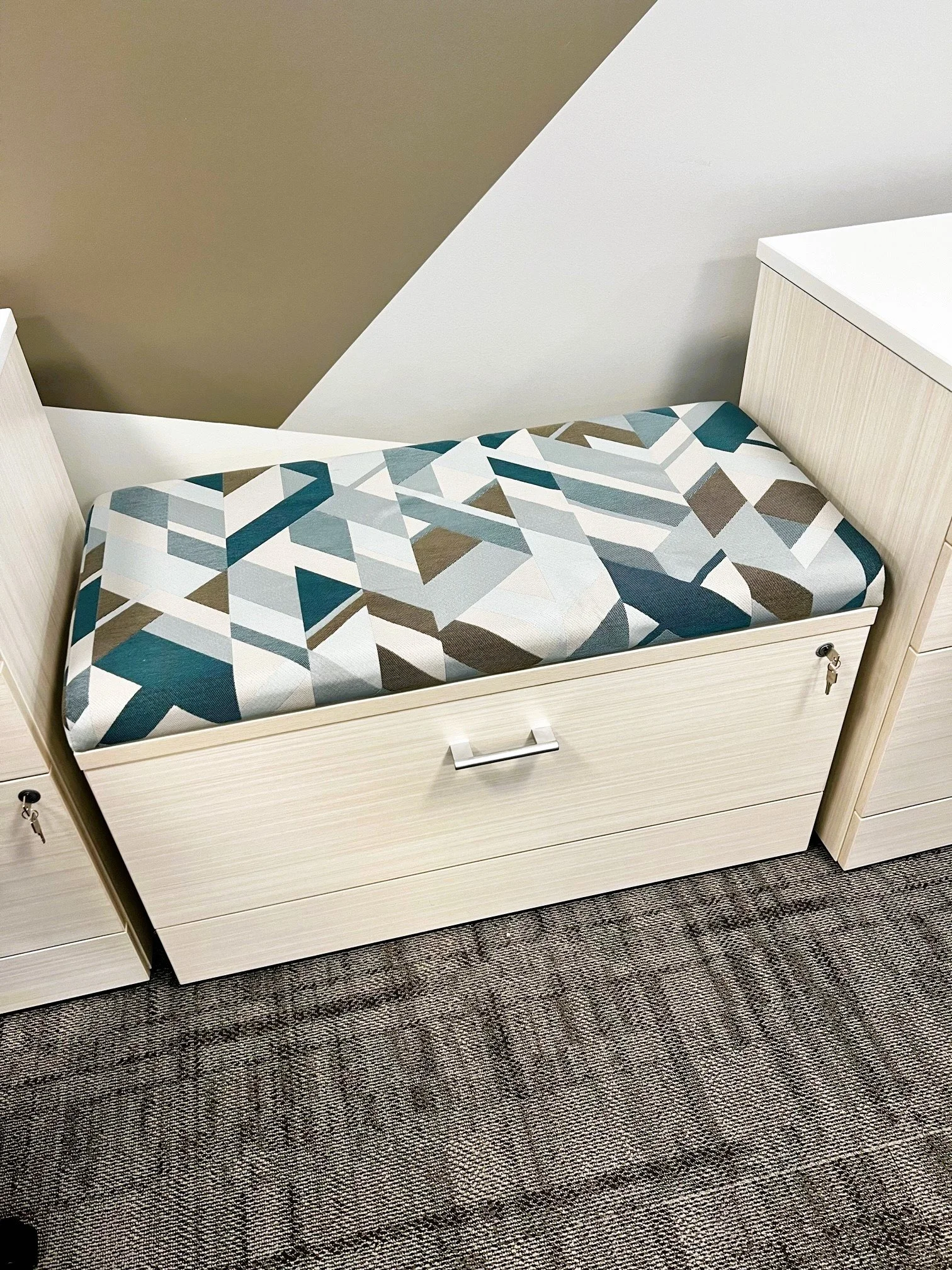 A beige-colored bench with a drawer, topped with a geometric patterned cushion, situated against a wall with a two-tone paint design.