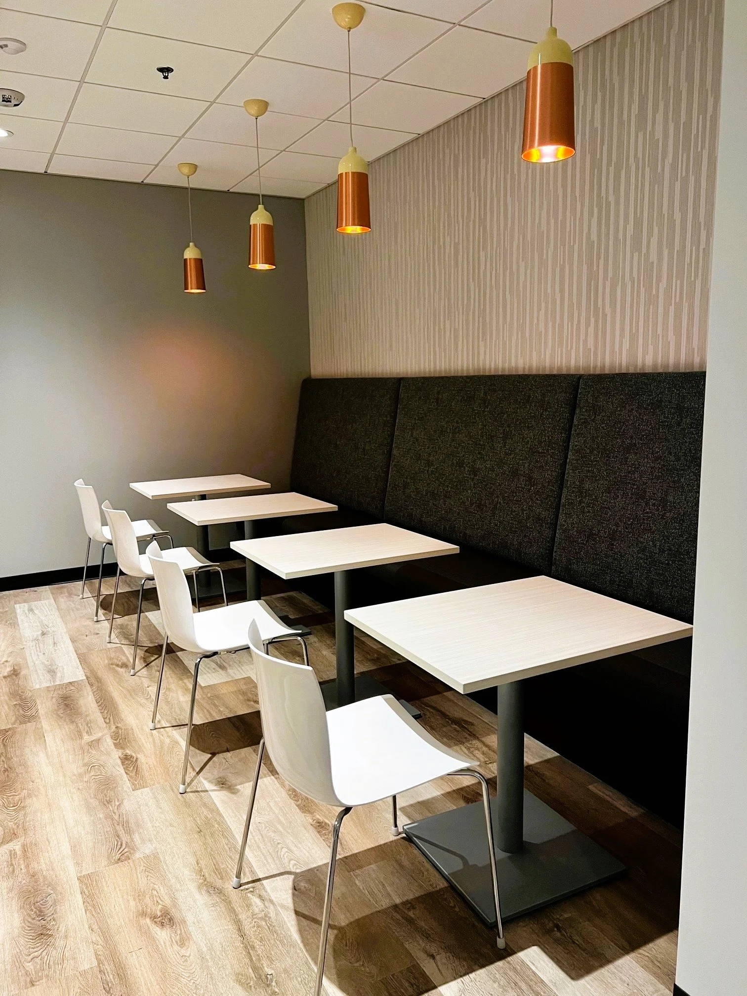 Empty seating area with four white chairs and four beige tables in a modern cafe with pendant lights and a textured wall.