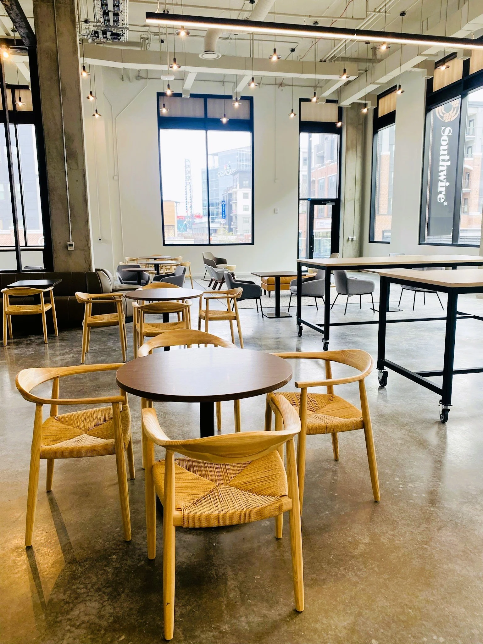 Modern, empty cafe interior with wooden and upholstered chairs, round and rectangular tables, large windows, concrete floor, exposed ceiling, and ambient hanging lights.