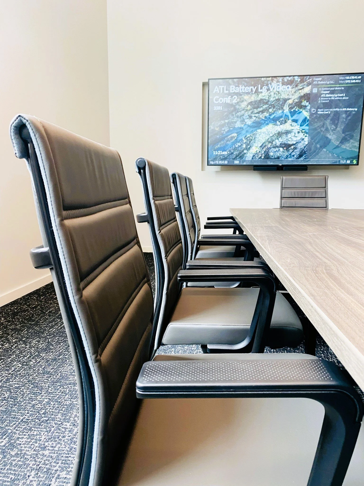 Empty conference room with six black and beige chairs aligned along a light wood table, with a wall-mounted TV displaying a battery-related video.