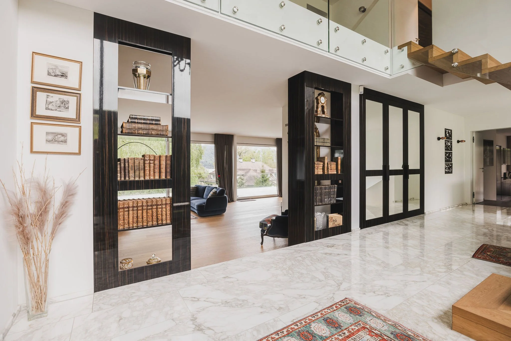 Innenaufnahme eines modernen Wohnzimmers mit Bücherregalen aus Ebenholz (Macassar), großen Fenstern, weißen Wänden, Marmorfußboden, einer schwarzen Couch im Hintergrund und einer Treppe aus Holz und Glas.