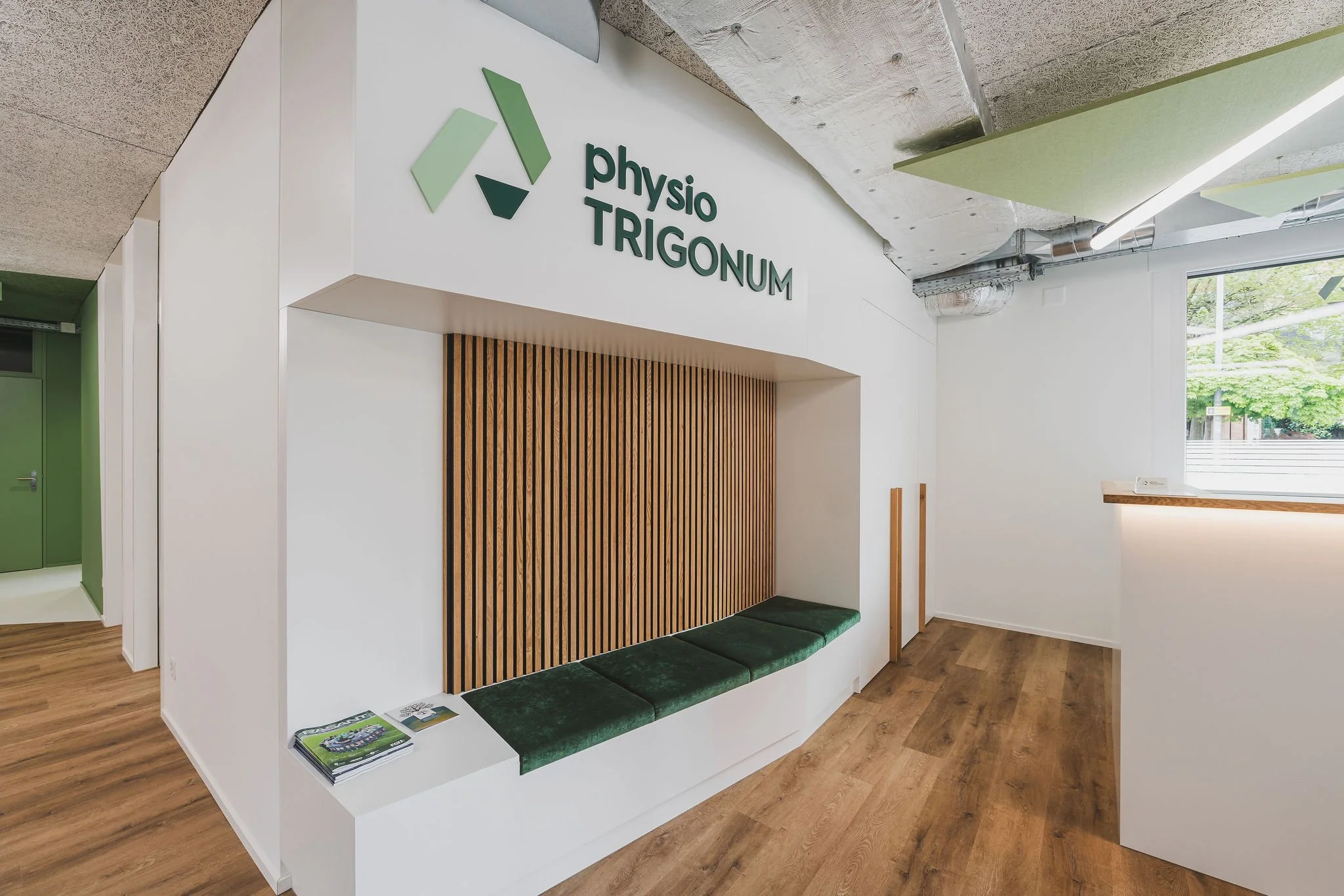Empfangsbereich mit stoffbezogene Sitzbank, Lamellenwand, Wandverkleidung, Schrankerweiterung und einem Logo mit der Aufschrift 'Physio Trigonom' an der Wand.