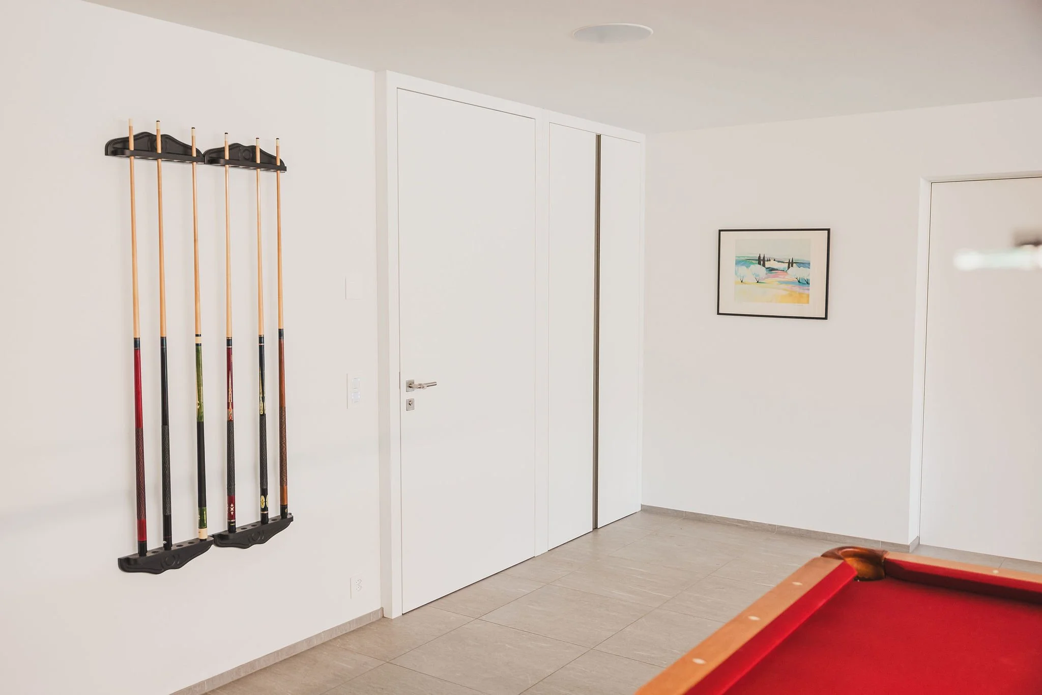 Ein Raum mit weißen Wänden, einer Tür mit Einbauschrank, einem Wandhalter mit Billardqueues, einer geahmten bunten Landschaftsmalerei und einem Billardtisch mit roter Felge.