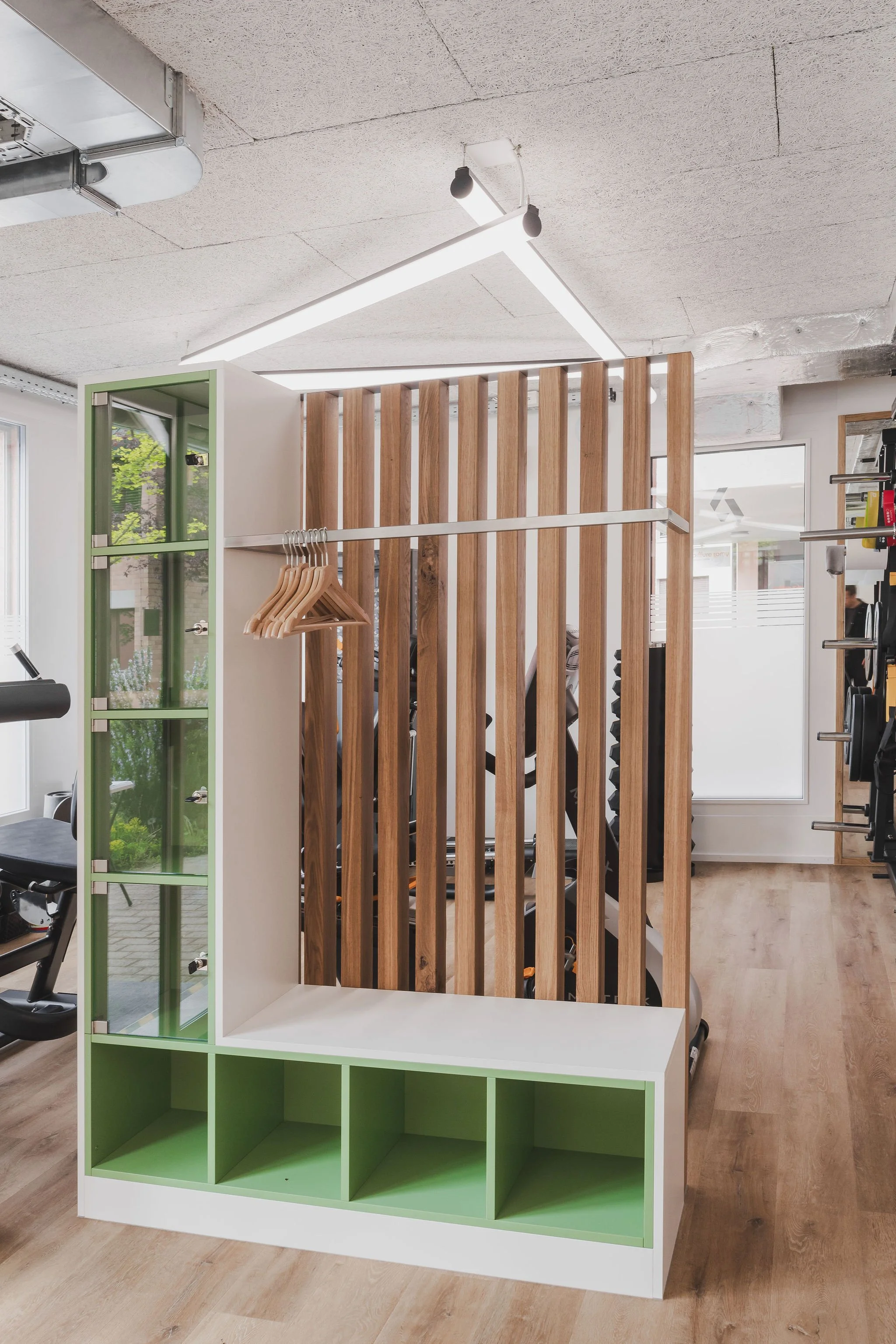 Innenaufnahme eines Fitnessstudios mit einer Holztrennwand, Garderobenregal mit Kleiderbügeln, einem Fitnessgerät und Gewichtsscheiben, helle Beleuchtung und große Fenster mit Blick nach draußen.