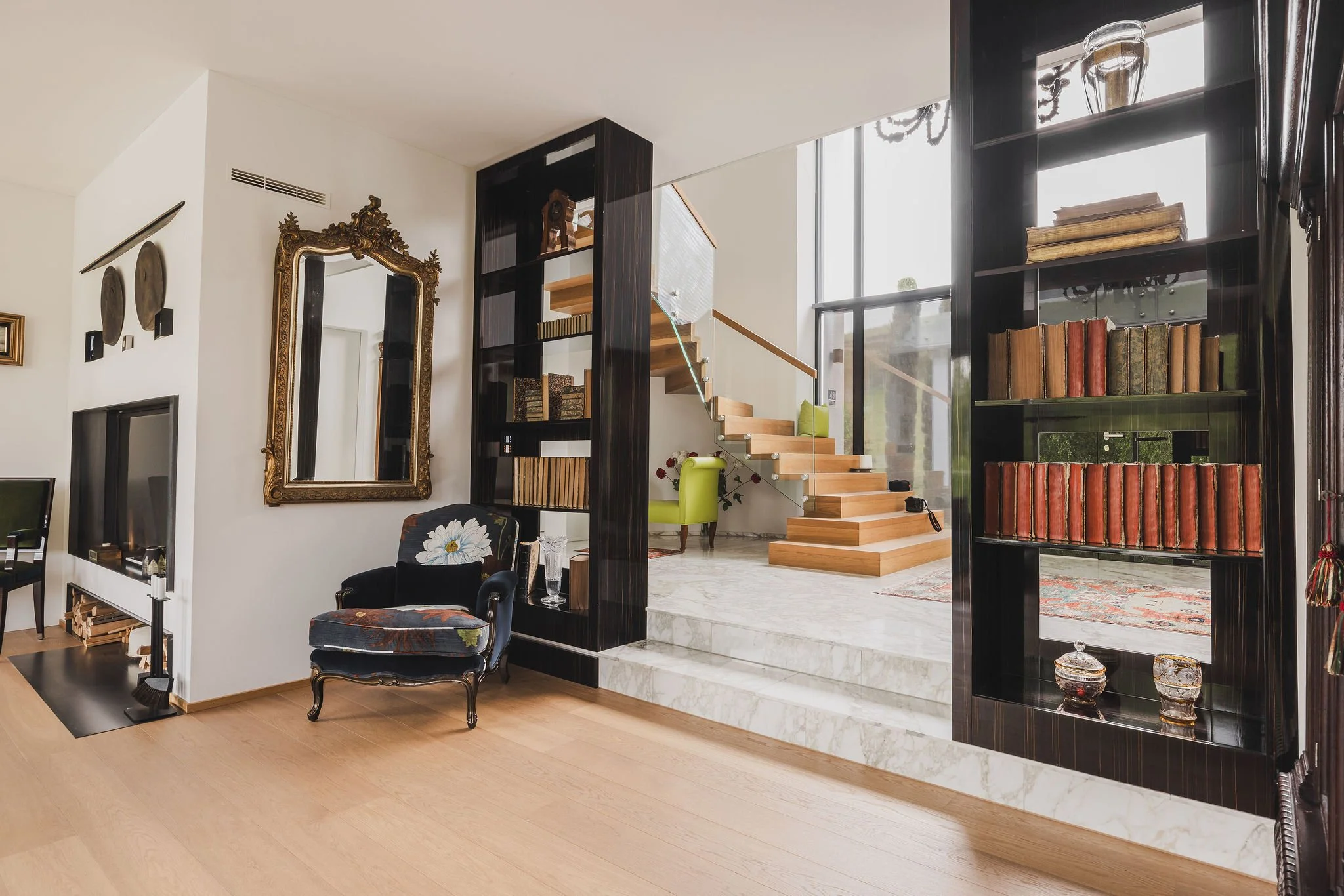 Ein Wohnzimmer mit einer schwarzen Bücherwand, einer schwarzen antiken Sessel mit einem Stoffmuster, einem großen goldenen Spiegel, einer Treppe mit Holzstufen, einer grünen Hausschuhe, einer Marokkanischen Teekanne auf der Regal und einem Fernsehger