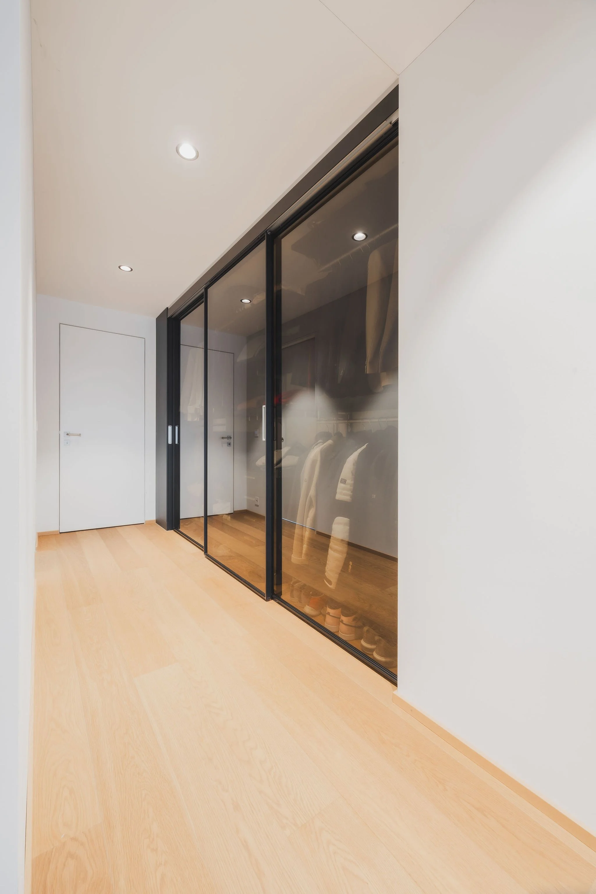 Ein Flur mit hellem Holzfußboden, weißer Wand und abgetrenntem begehbarem Kleiderschrank mit schwarzen Glasschiebetüren von Rimadesio. Im Schrank sind Jacken und Schuhe sichtbar.