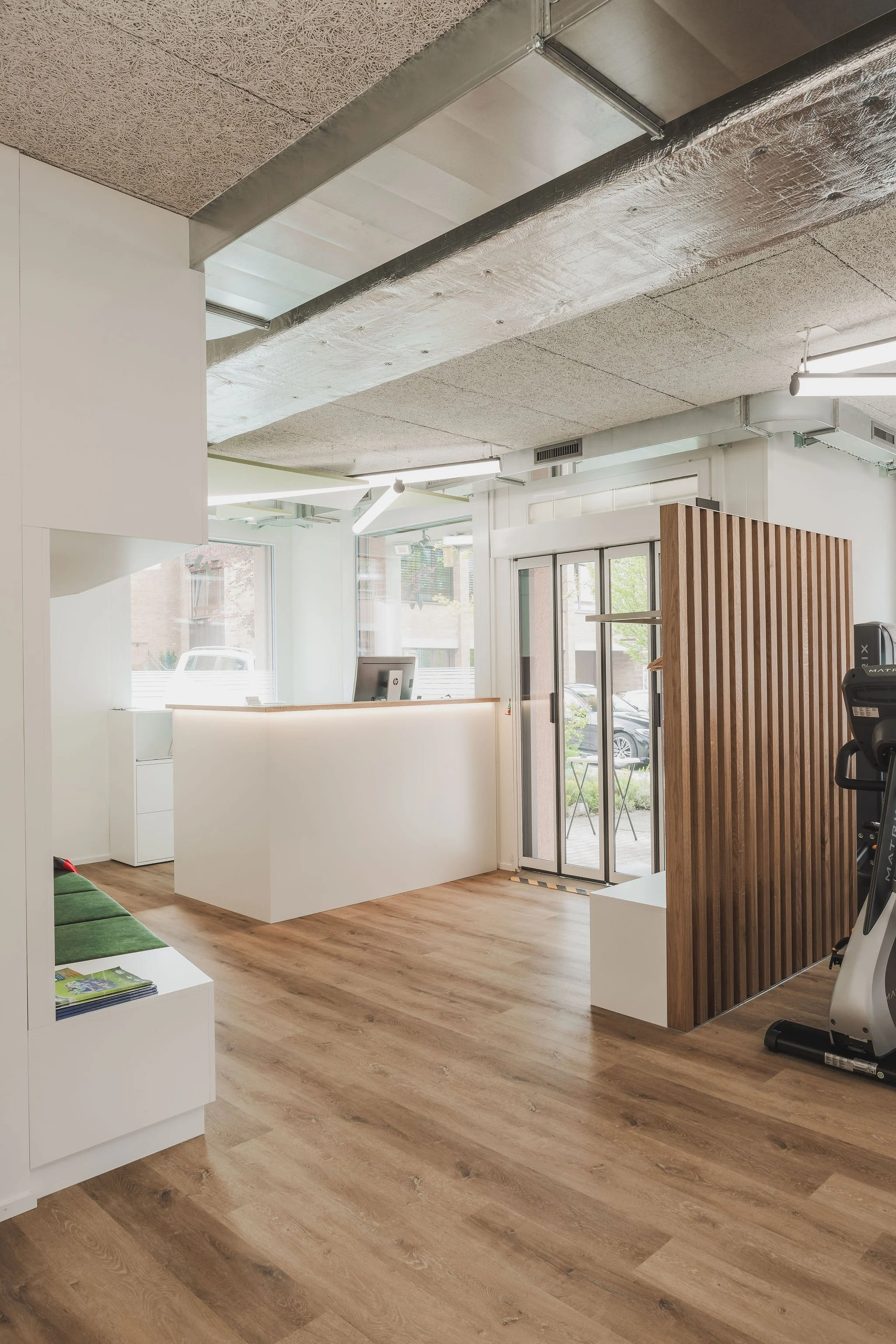 Innenraum eines modernen, hellen Wohn- oder Geschäftsraums mit Holzfußboden, weißen Wänden, Empfangstresen, einer Holzverkleidung und Fitnessgeräten.