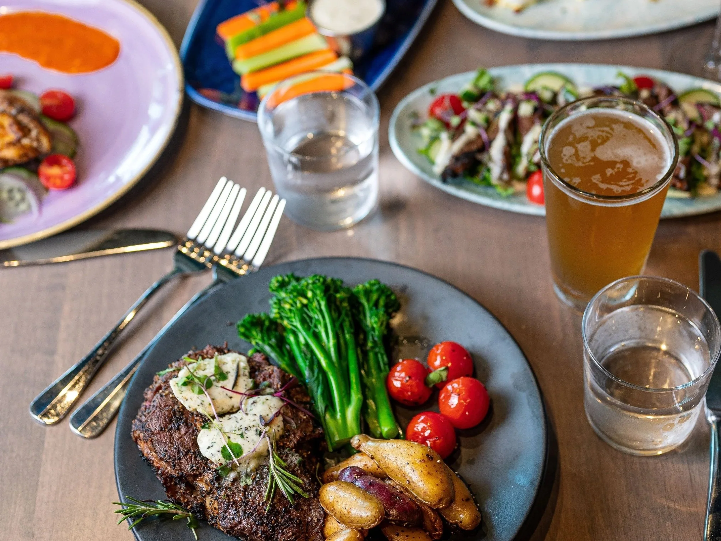 Overhead view of a dining table with plated steak, vegetables, appetizers, beer, cocktails, and shared dishes arranged for a group meal.