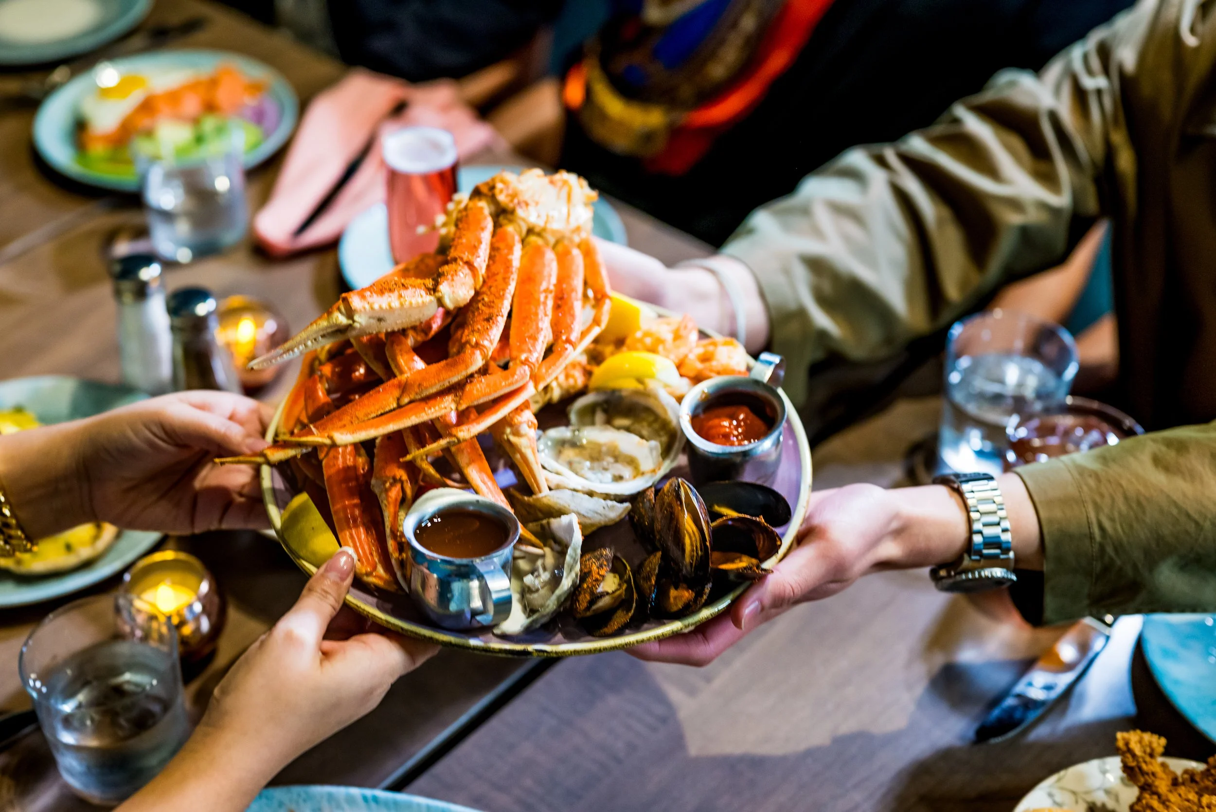 Hands pass a large seafood platter across a dining table set with drinks, candles, and shared dishes, including crab legs, mussels, shrimp, lemon wedges, and dipping sauces.