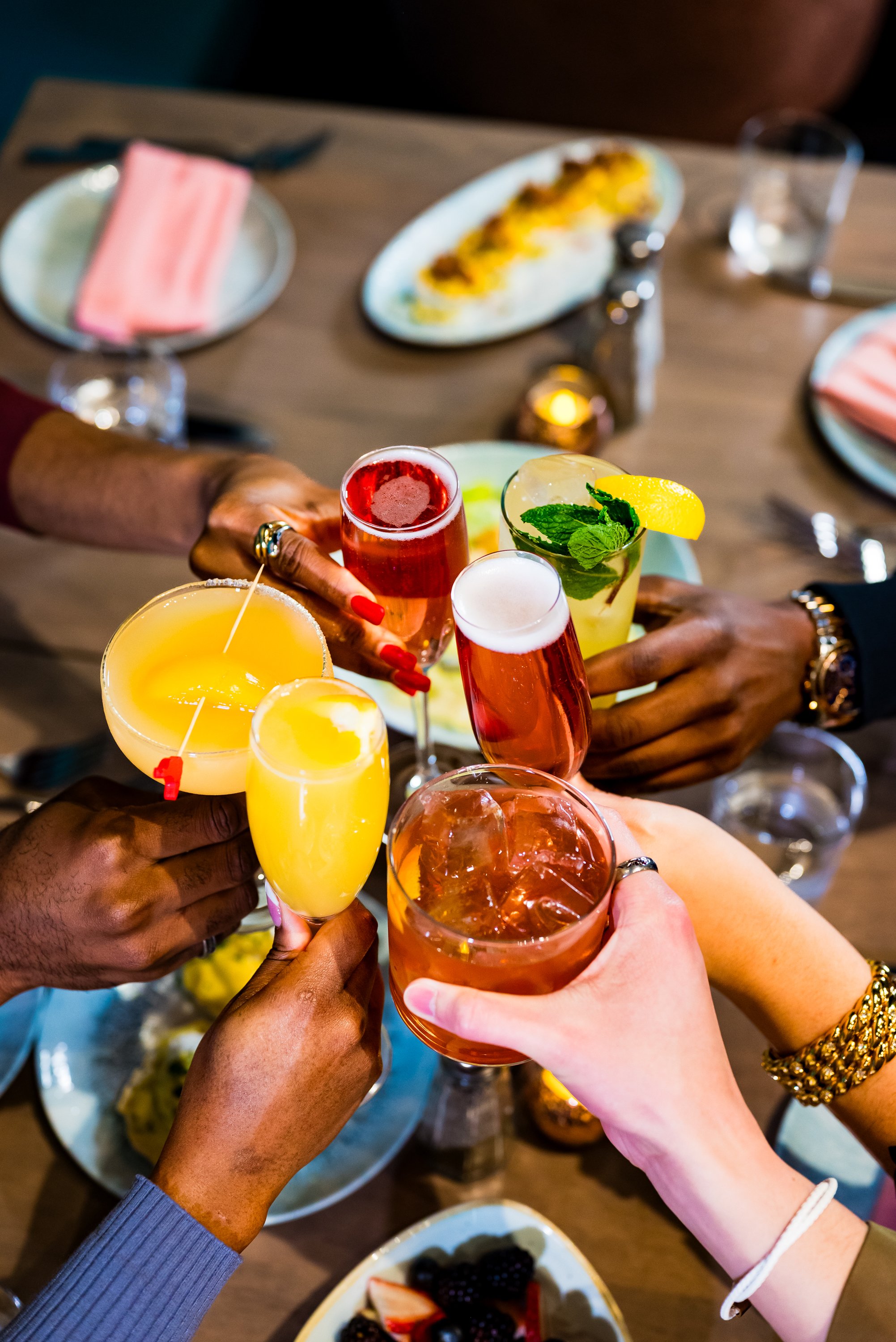 A group of people raising colorful cocktails over a dining table set with plates of food, berries, a rice dish, utensils, napkins, and a lit candle during a celebratory meal.
