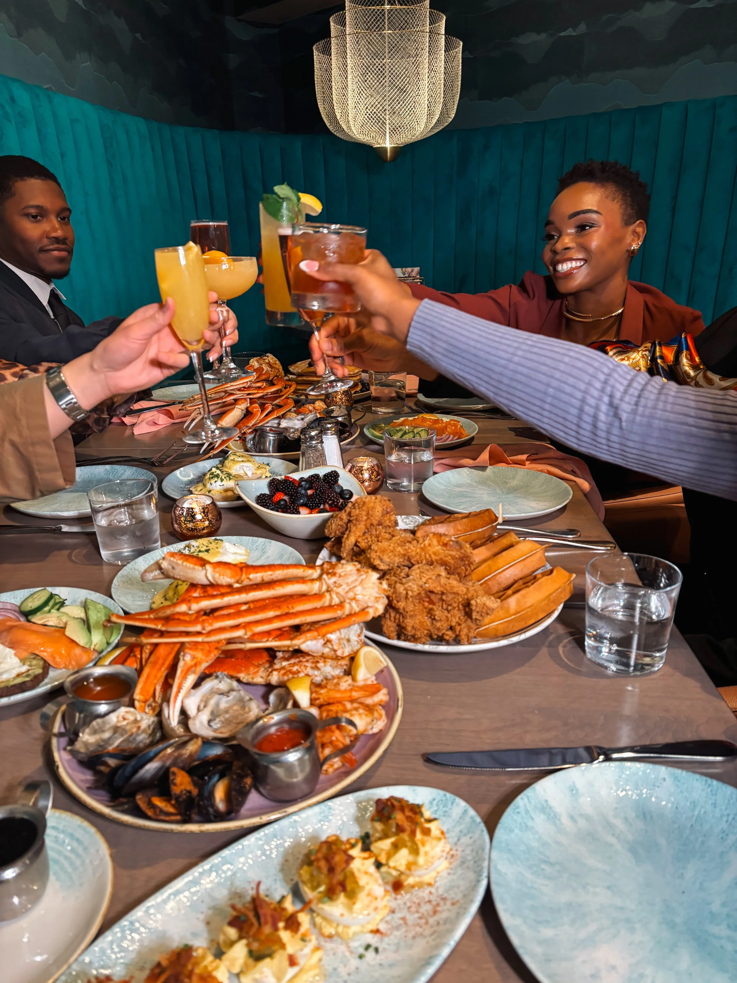 A table filled with seafood dishes, fried items, deviled eggs, and drinks, with several people raising glasses for a toast in a stylish restaurant setting.