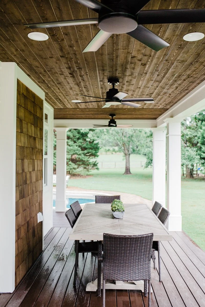 Outdoor patio with wooden ceiling, ceiling fans, and wicker dining set on wooden deck.