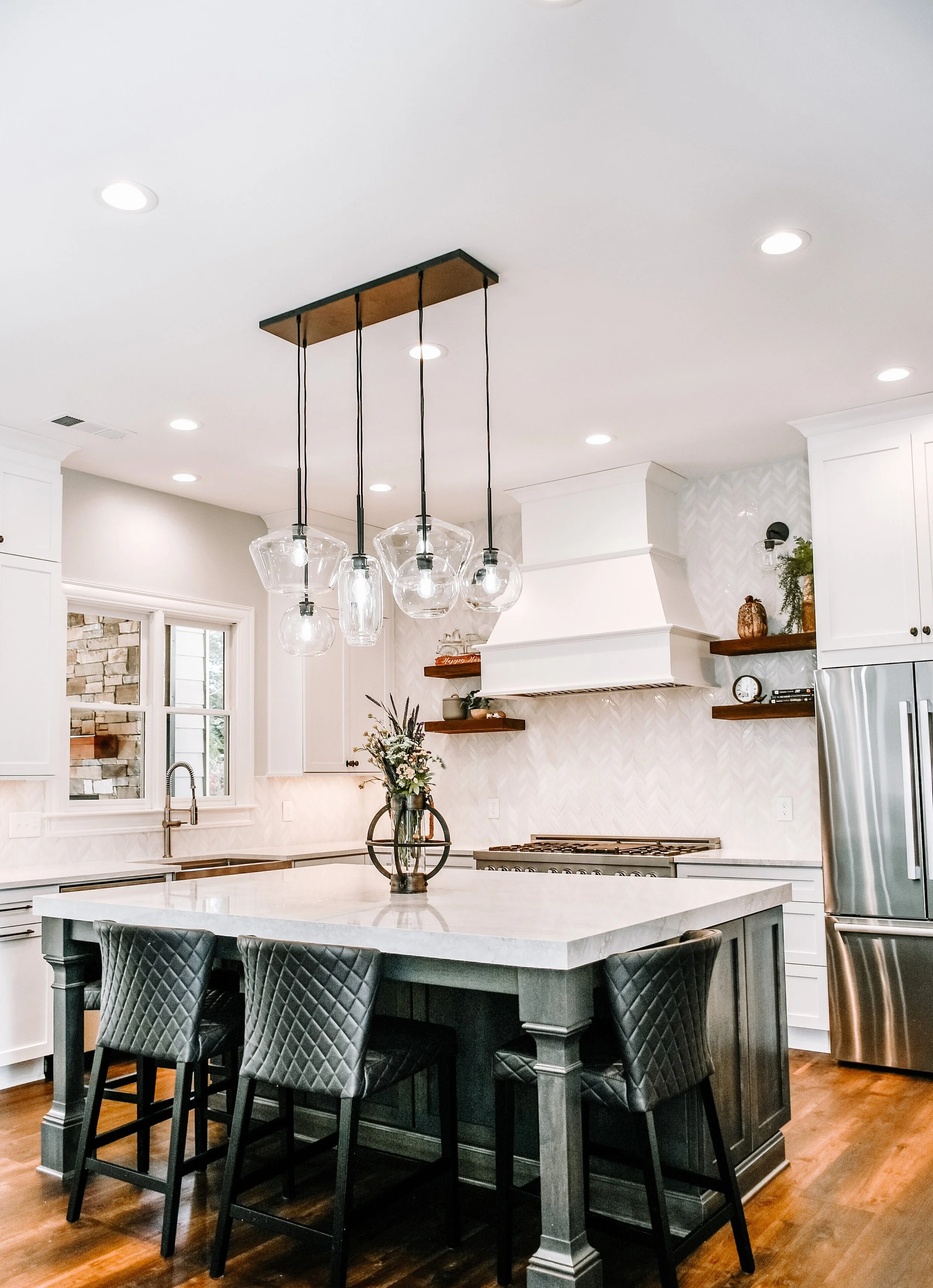 Modern kitchen with pendant lights, island, bar stools, white cabinetry, marble countertops, stainless steel appliances, wooden shelves, and floral arrangement.
