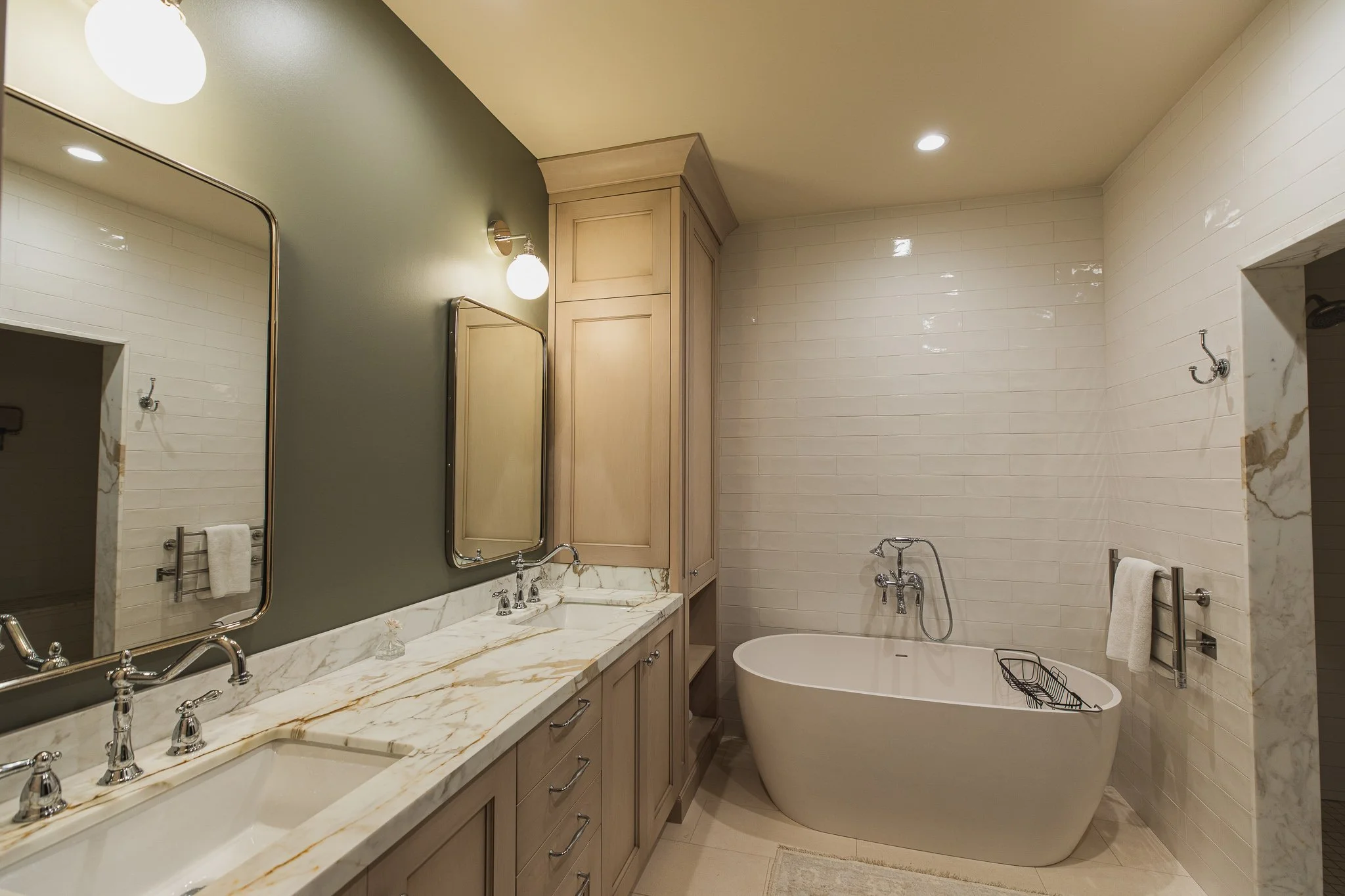 Modern bathroom with double sinks, marble countertop, freestanding bathtub, wall-mounted faucets, green accent wall, and wooden cabinetry.