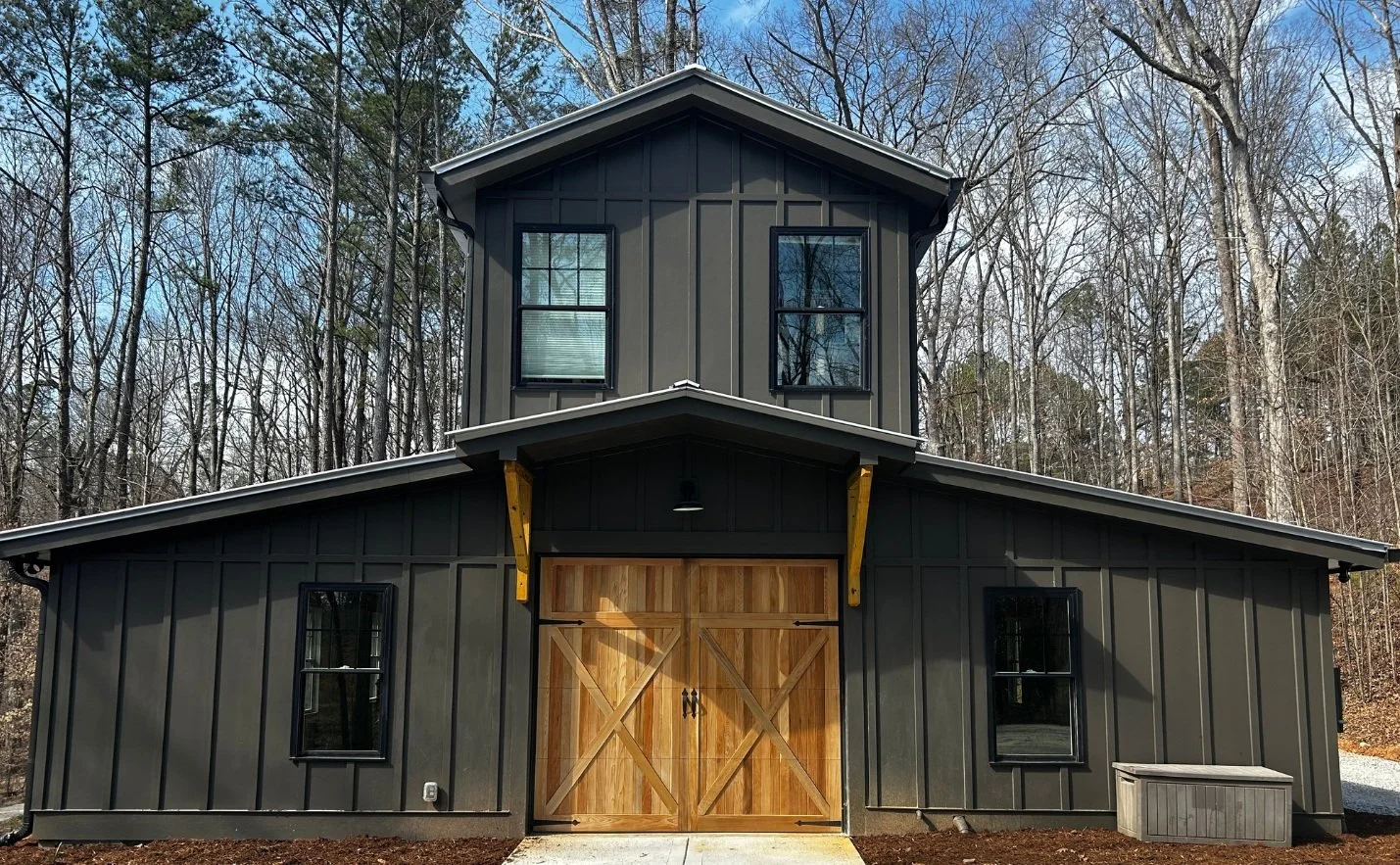 Two-story black barn-style building with wooden double doors, black framed windows, and surrounded by bare trees.