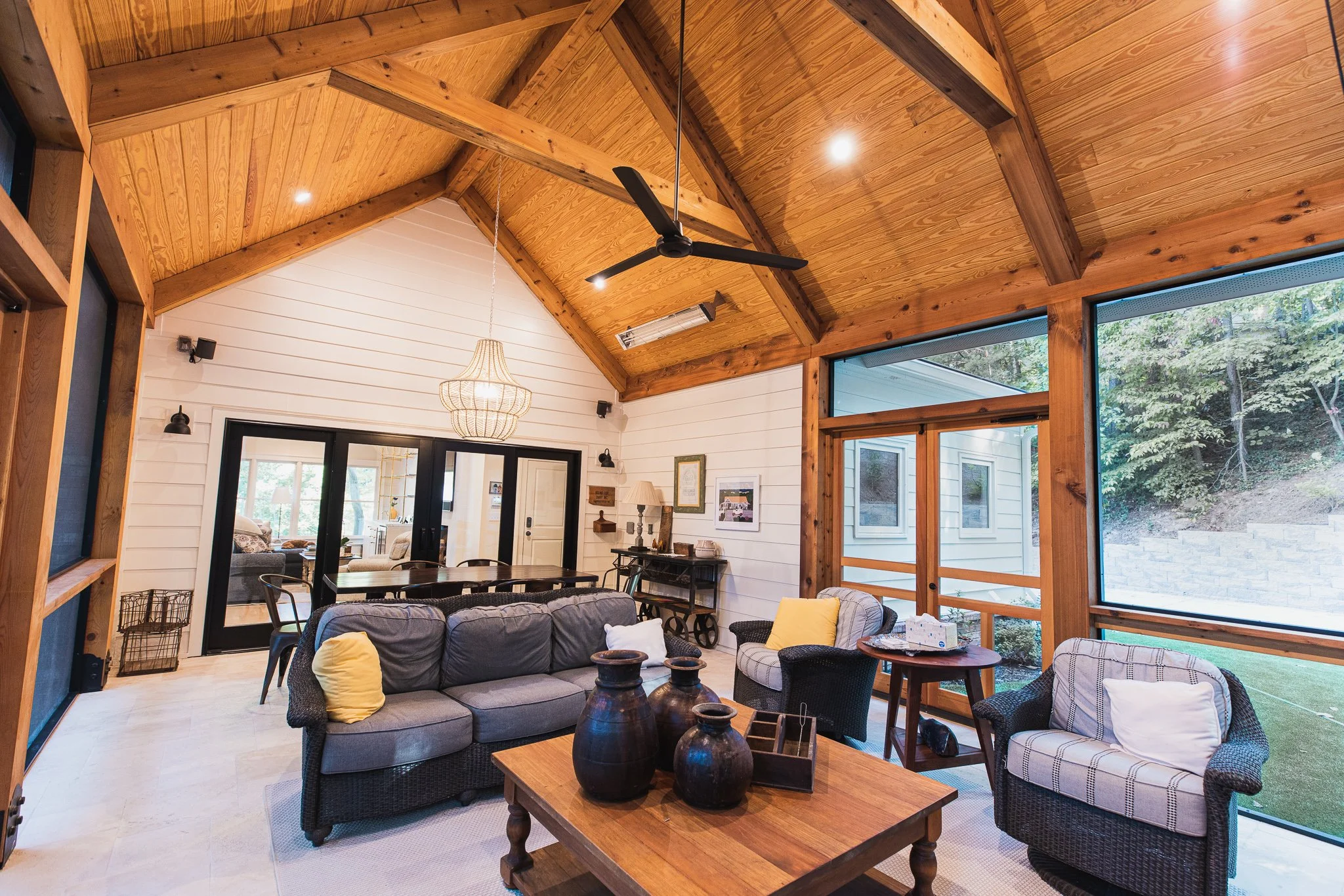 Spacious room with vaulted wooden ceiling and large windows, featuring wicker furniture, a coffee table with vases, a dining table, and a ceiling fan. The room is decorated in a cozy style with earthy tones.