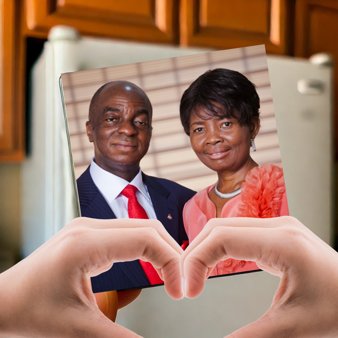 A hand holding a heart-shaped frame around a photograph of a man in a suit and woman in a pink outfit, with a kitchen in the background.