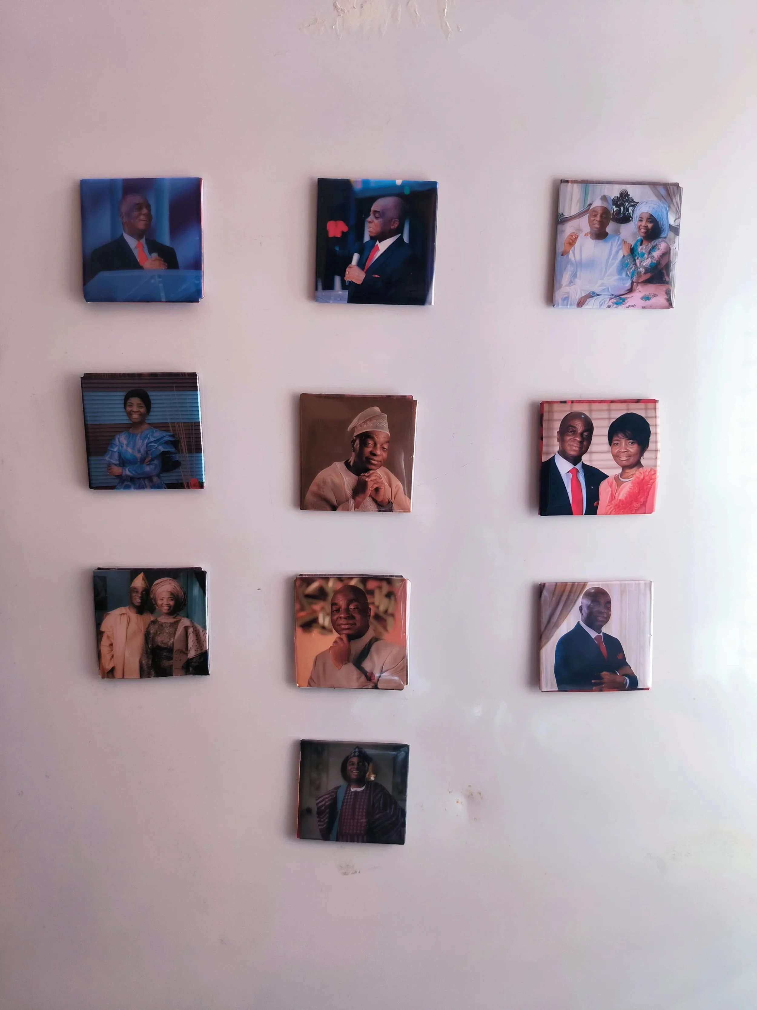 A wall decorated with eight framed photographs of people, arranged in a grid pattern with three columns and four rows. The photos include various individuals, some dressed in traditional Nigerian attire and others in Western clothing.
