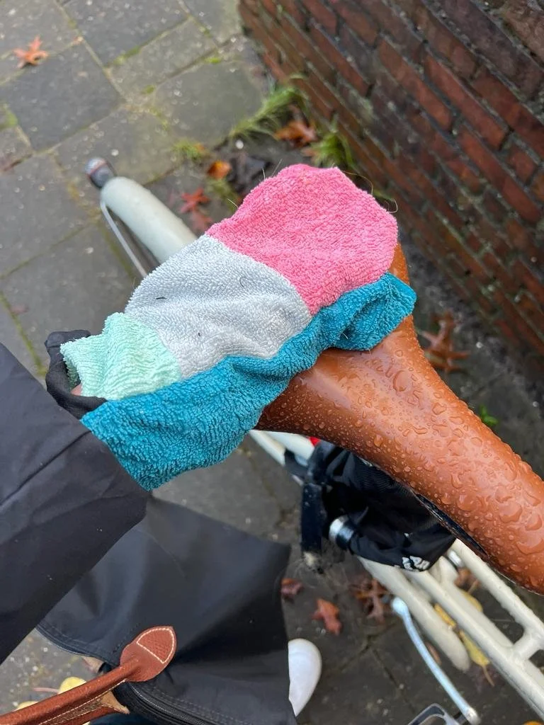 Close-up of a person's hand wearing a multicolored tobi towel glove, covering the bike saddle which has droplets of water on it, on a wet sidewalk with fallen leaves.