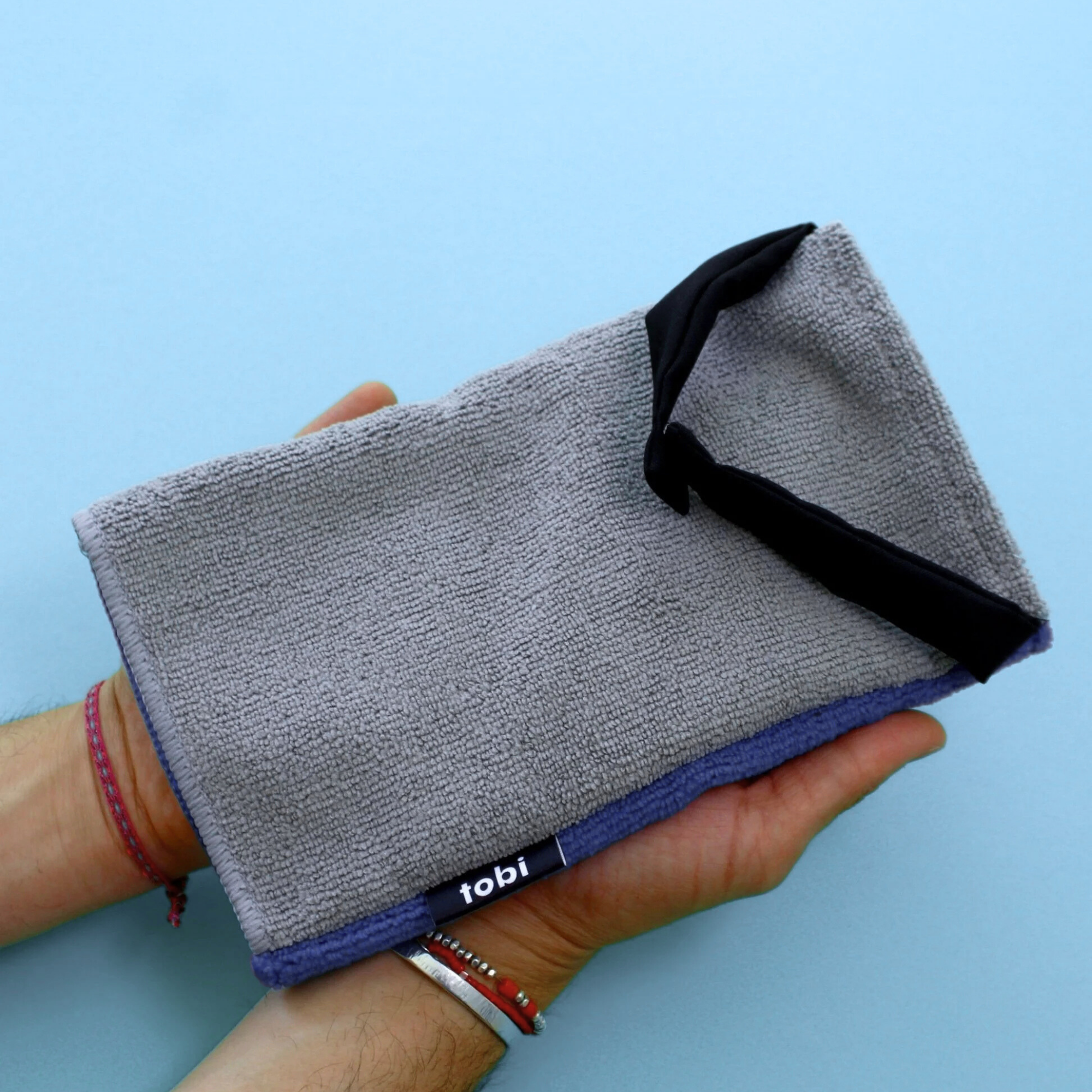 A hand holds a grey tobi towel with two black ends, with a small black label that reads 'tobi,' against a light blue background.