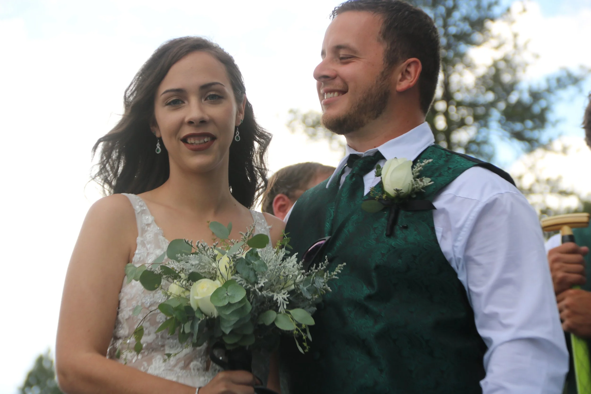 Bride and groom standing outdoors during wedding ceremony, with the bride holding a bouquet of white roses and greenery, both smiling and dressed in formal wedding attire.