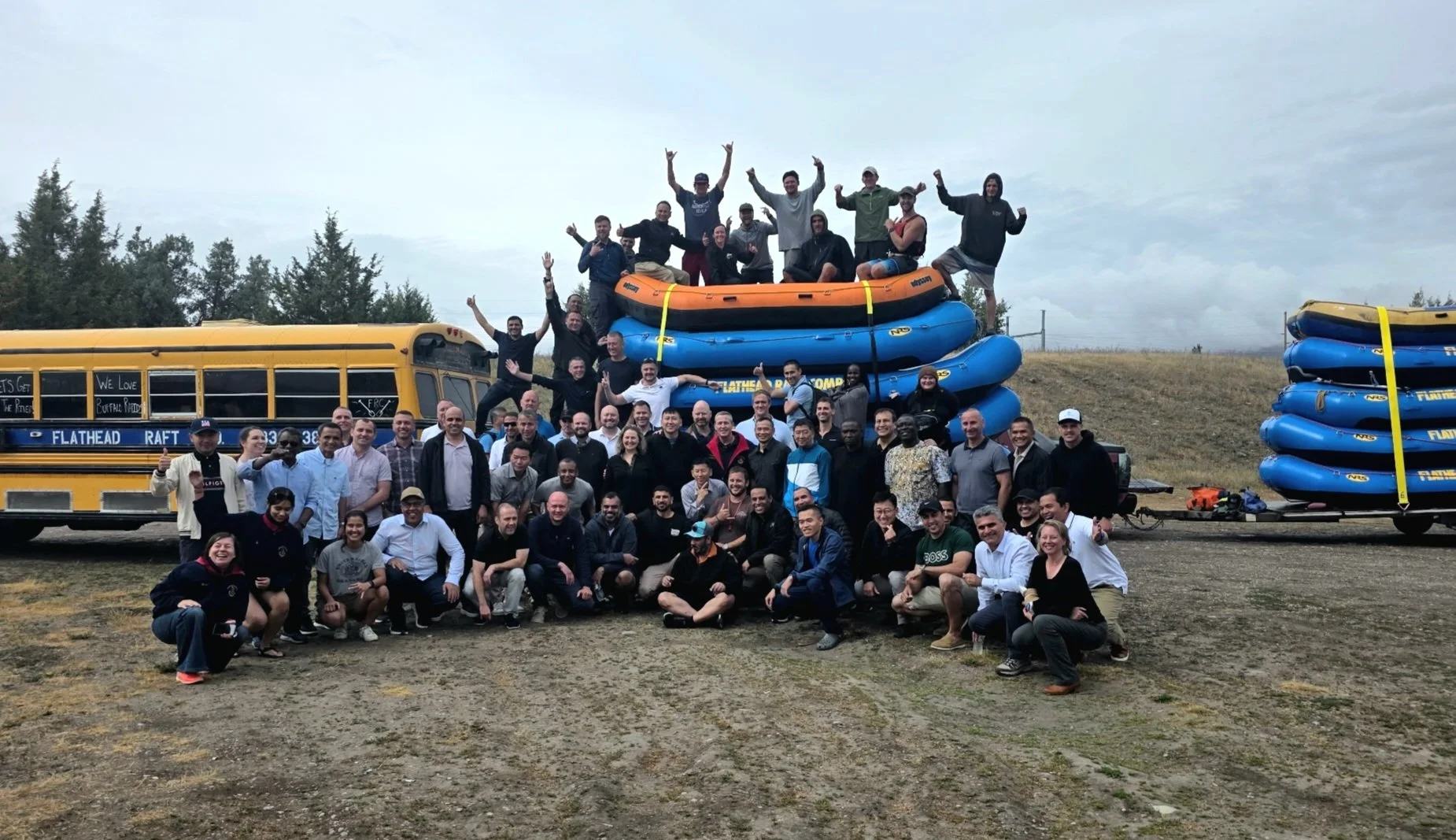 Group of people gathered around and on top of outdoor rafting equipment, including stacked inflatable rafts, with a yellow school bus in the background, under cloudy skies.