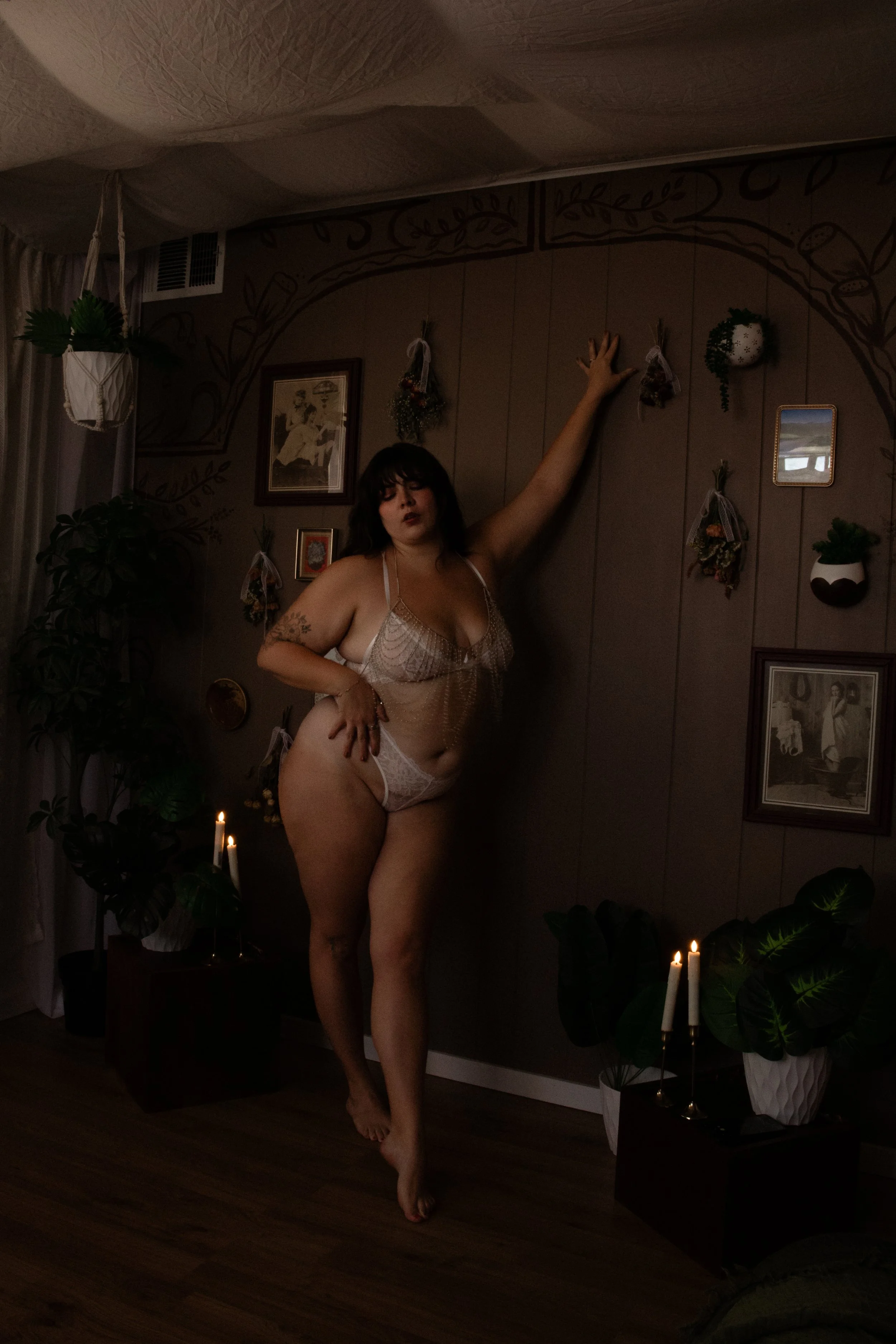 A woman posing in a dimly lit room wearing lace lingerie, surrounded by plants and candles, with wall decor including framed pictures and hanging arrangements.