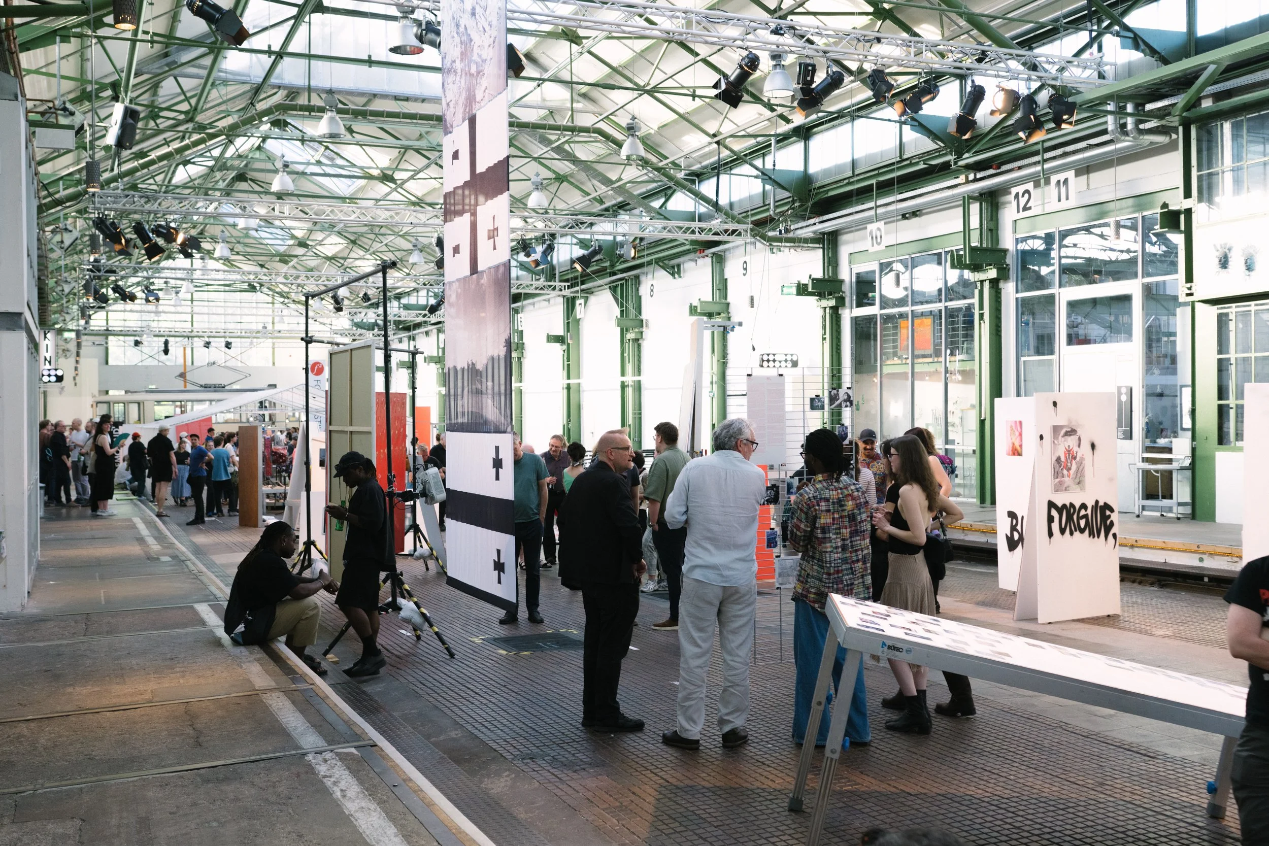 An indoor art exhibition with people viewing artwork and engaging in conversations, features high ceilings with natural light and hanging lights, and large glass doors in the background.