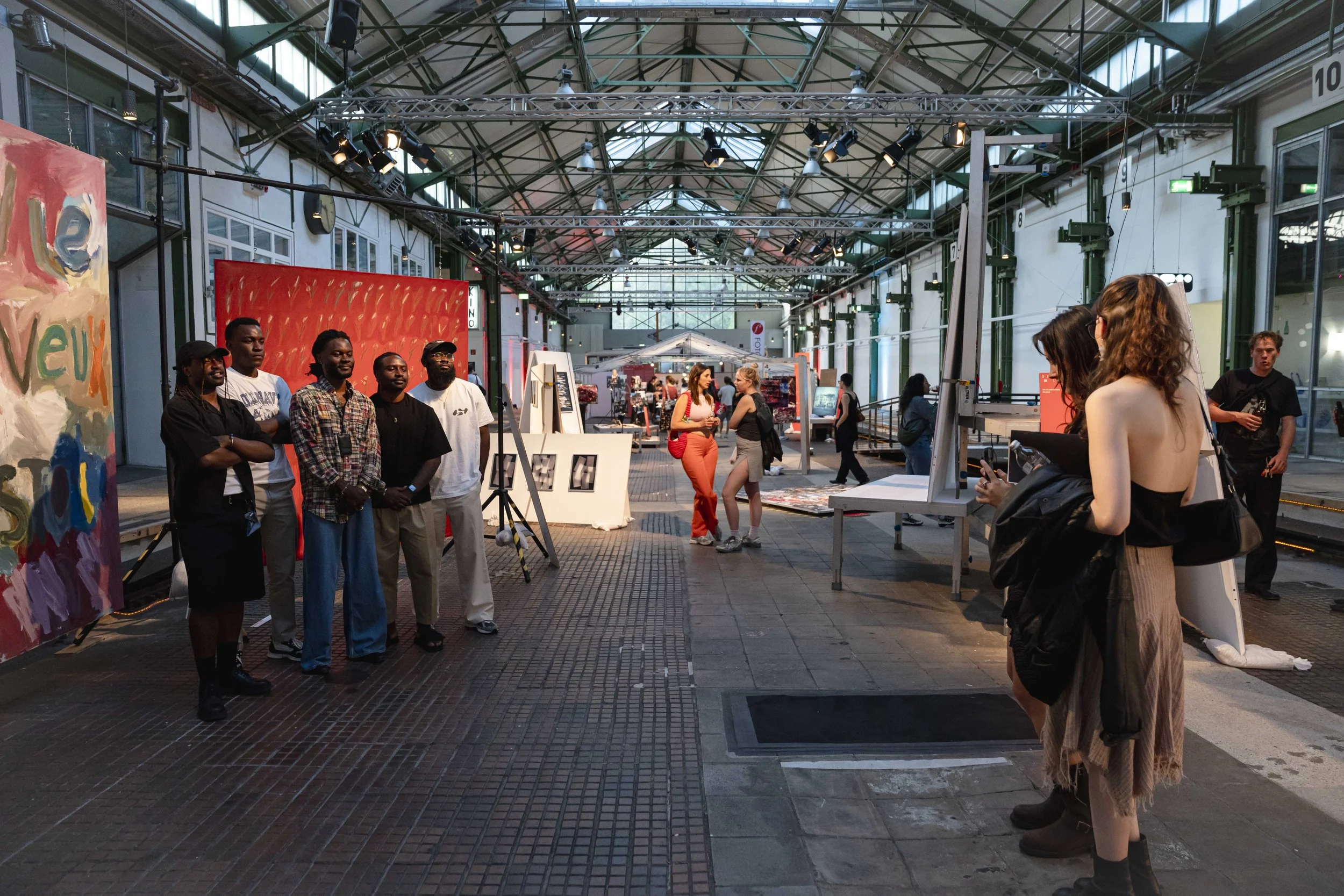 People at an art exhibition inside a spacious, glass-roofed industrial-style gallery with various artworks on display.