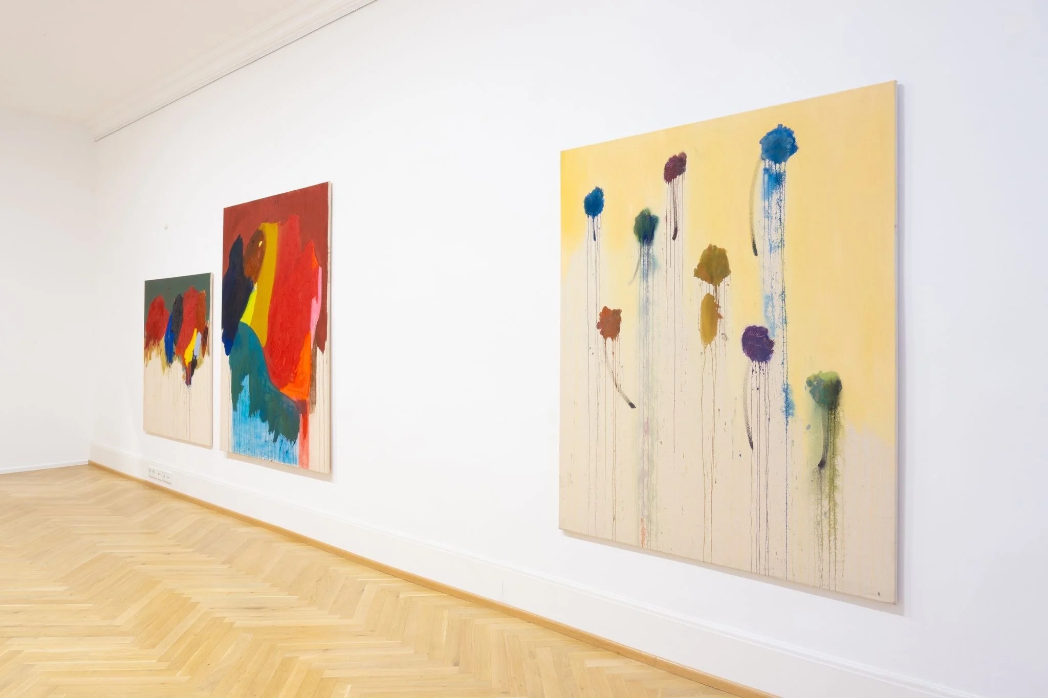 Abstract paintings with colorful shapes and drips hanging on a white gallery wall with a wooden floor.