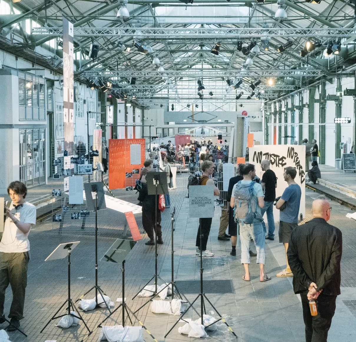 A crowded indoor exhibit space with people observing various displays and signs, some with protest messages like 'NO JUSTICE NO PEACE'. The area has industrial architecture with metal framework and large windows, and there are musical stands and equipment in the foreground.
